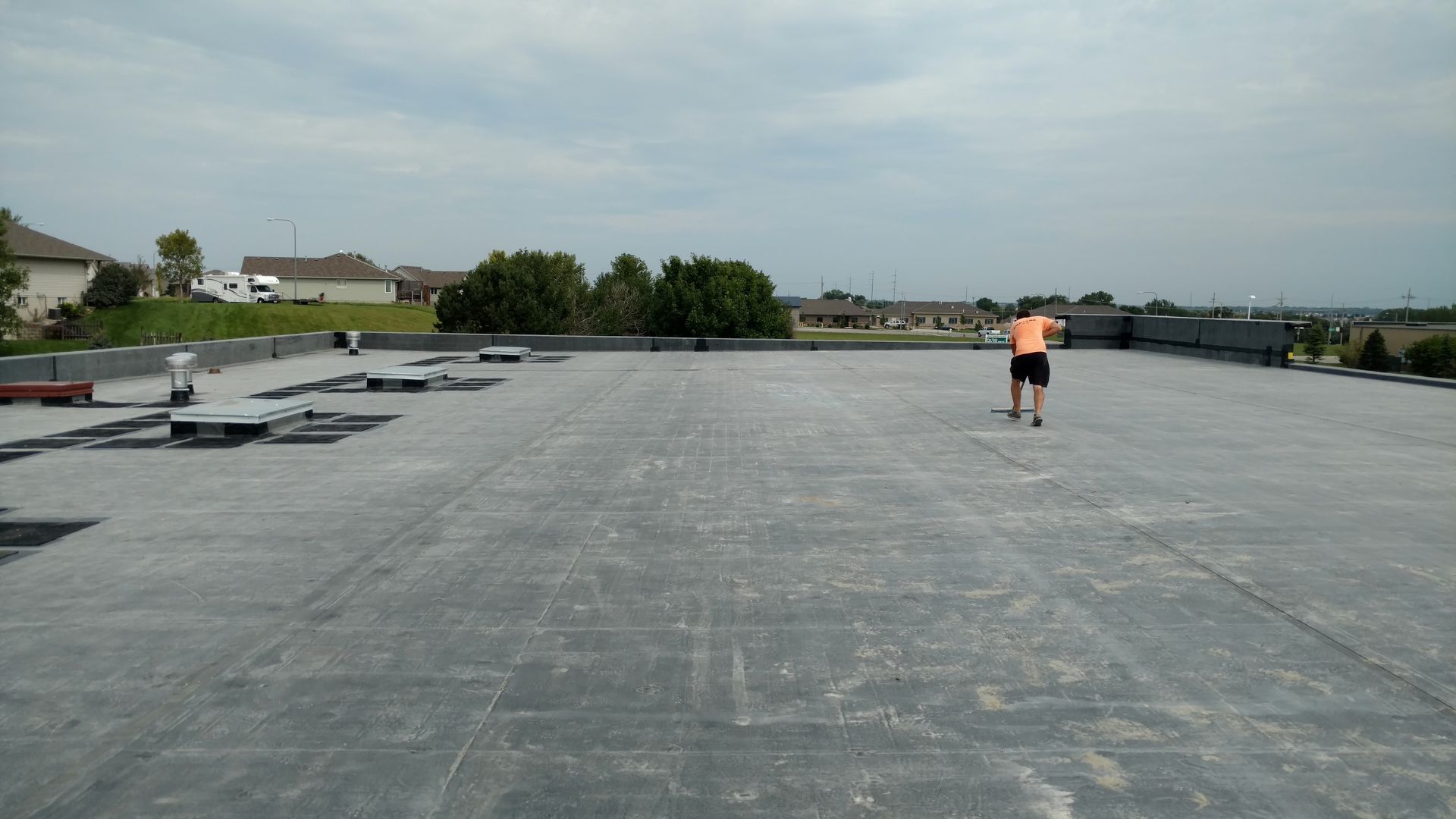 A person in a bright orange shirt walks across a large, flat gray commercial roof under a cloudy sky.