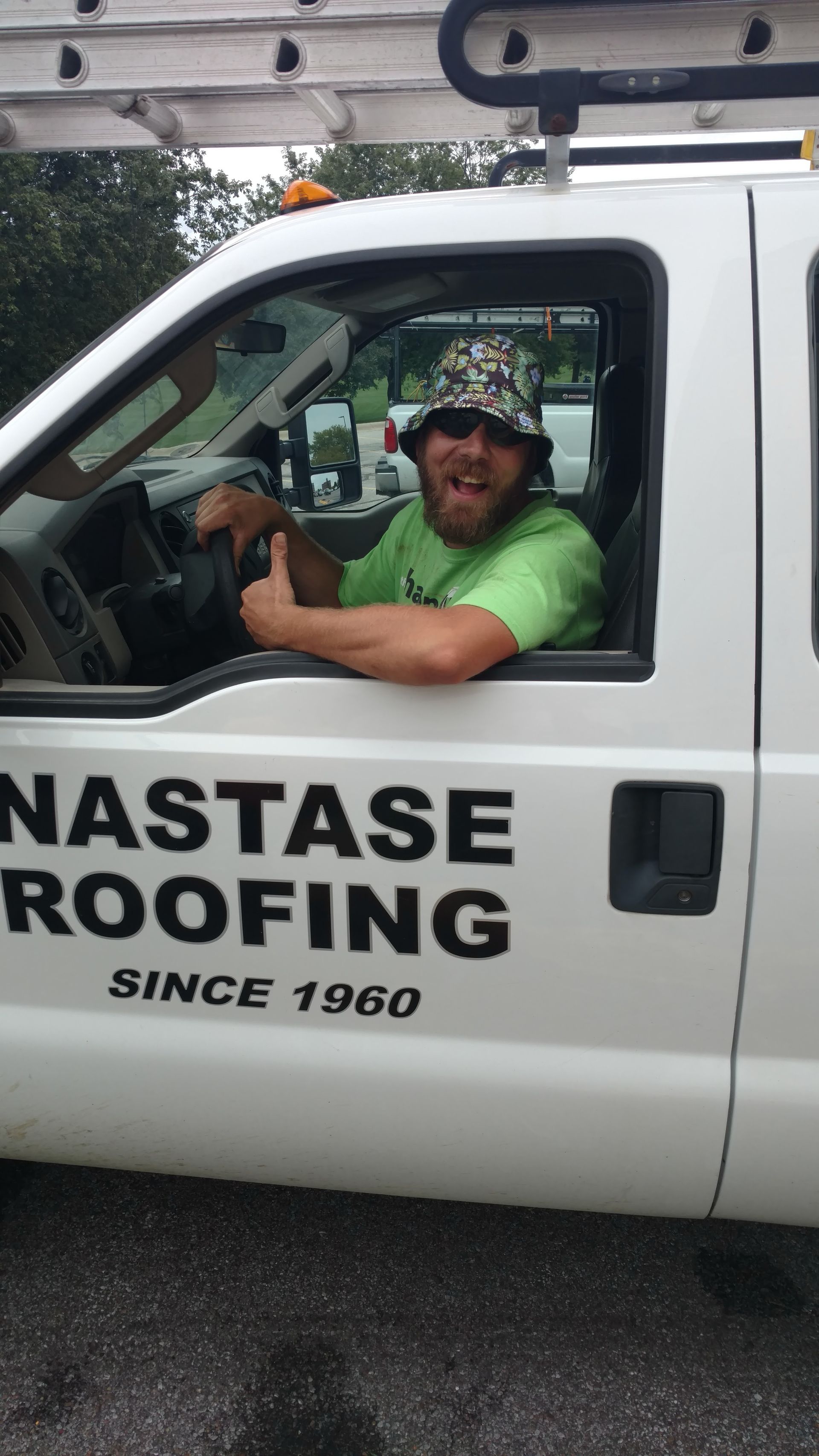 A person in a bucket hat and green shirt smiles while sitting in the driver's seat of a white Nastase Roofing work truck.