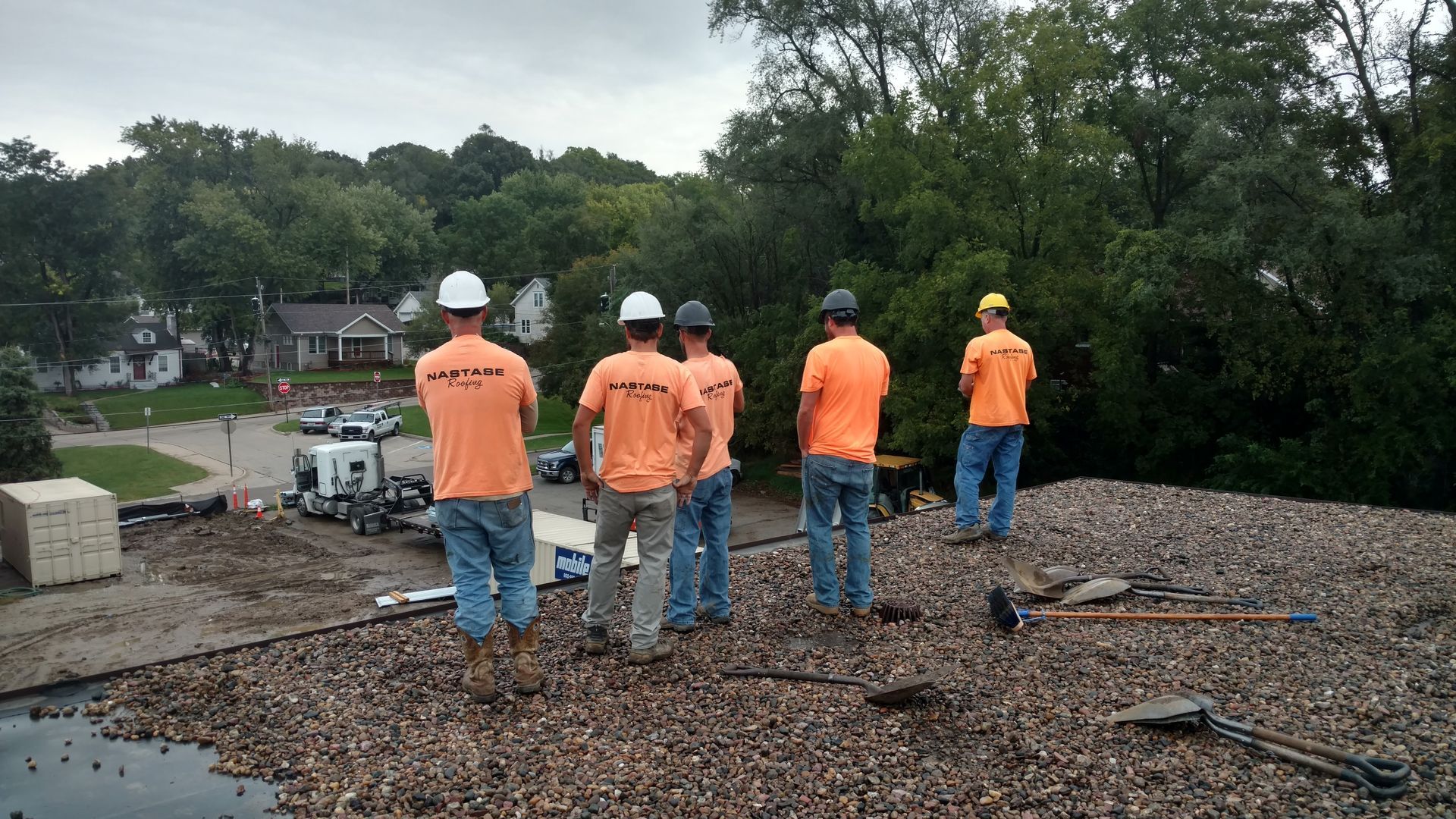 Five workers in orange shirts and hard hats stand on a gravel roof, looking out toward a residential street and trees.