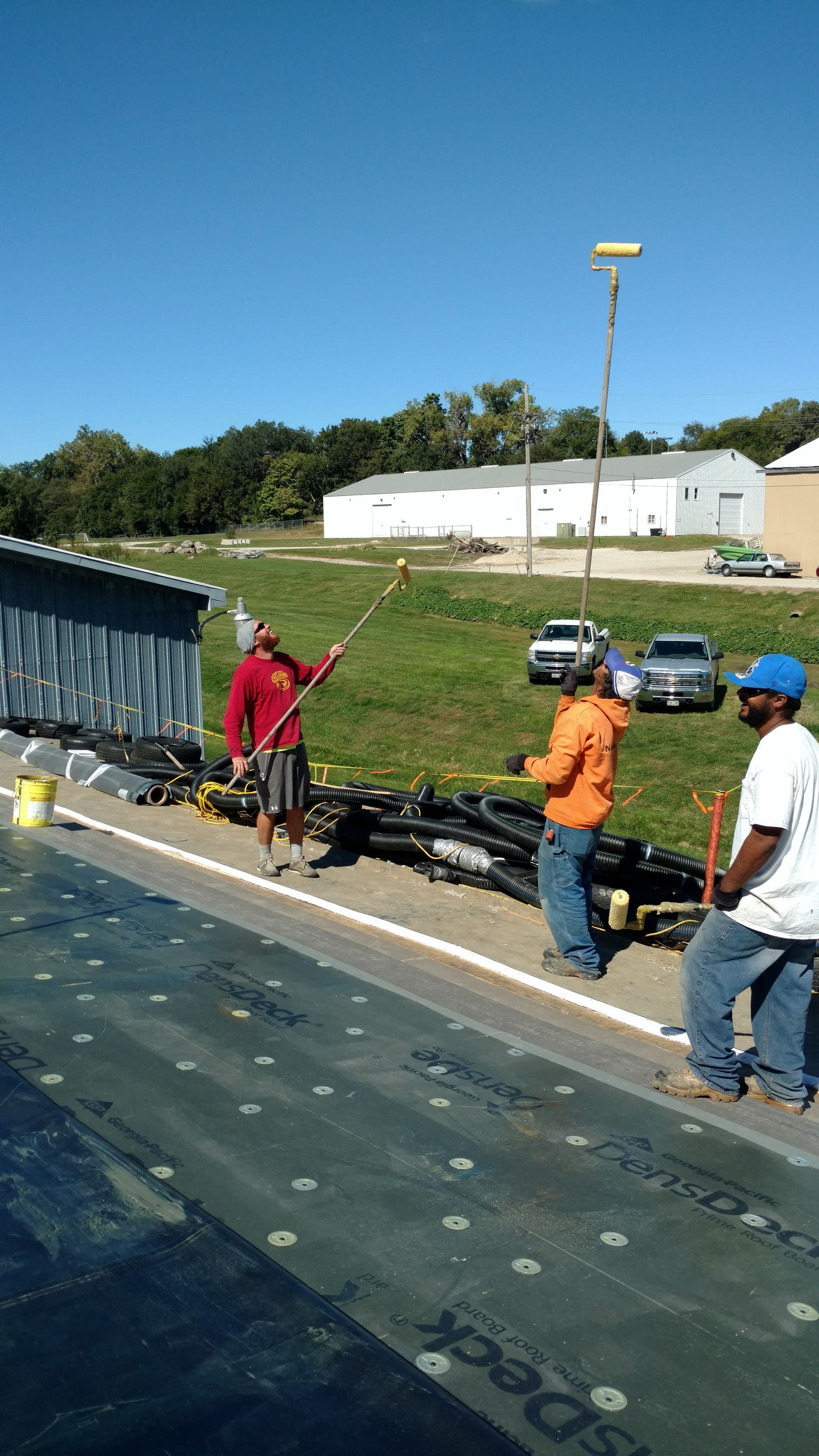 Three construction workers on a rooftop install materials on a sunny day near a field and parking lot.