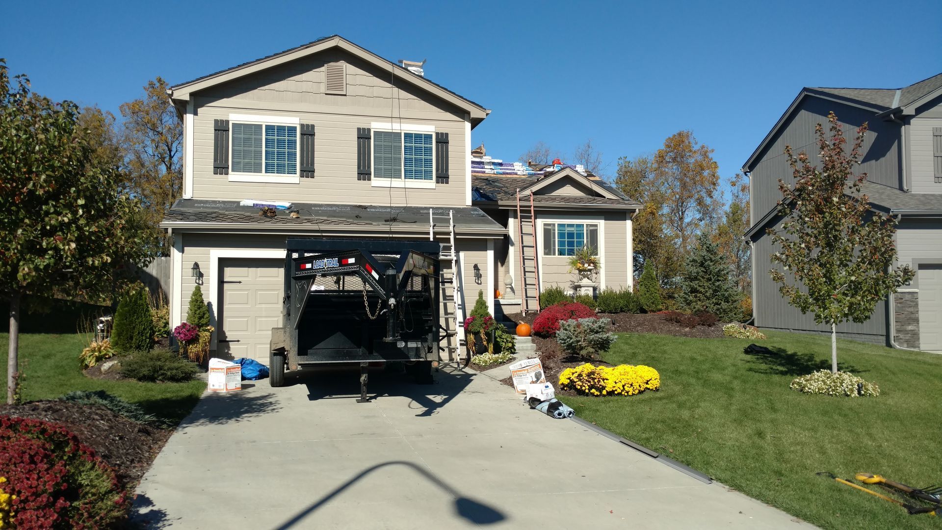 Two-story suburban house with a trailer in the driveway and workers on the roof during a daytime repair project.