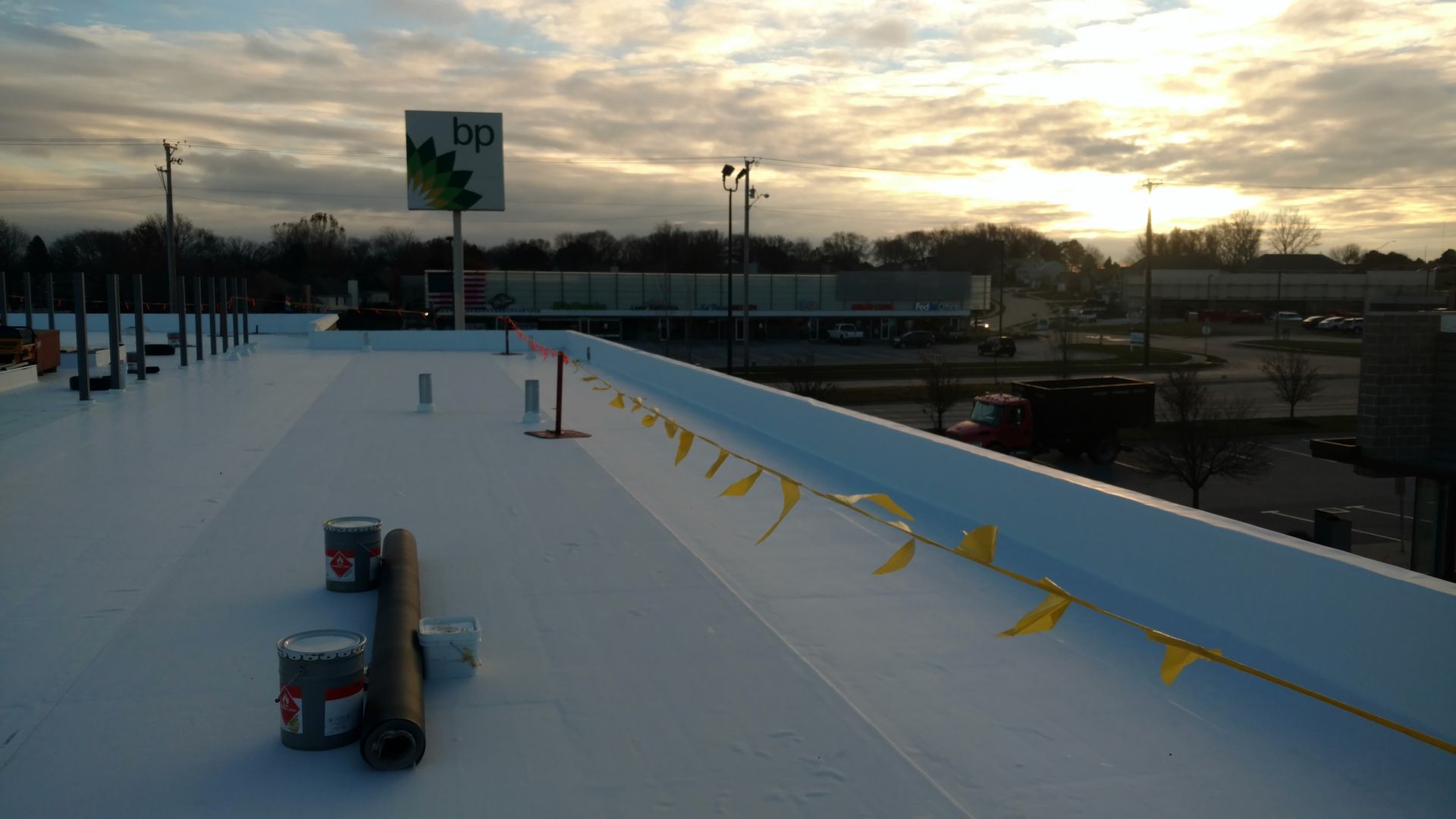 A rooftop under construction with white membrane, a strip of yellow flags, and materials at sunset near a BP gas station.