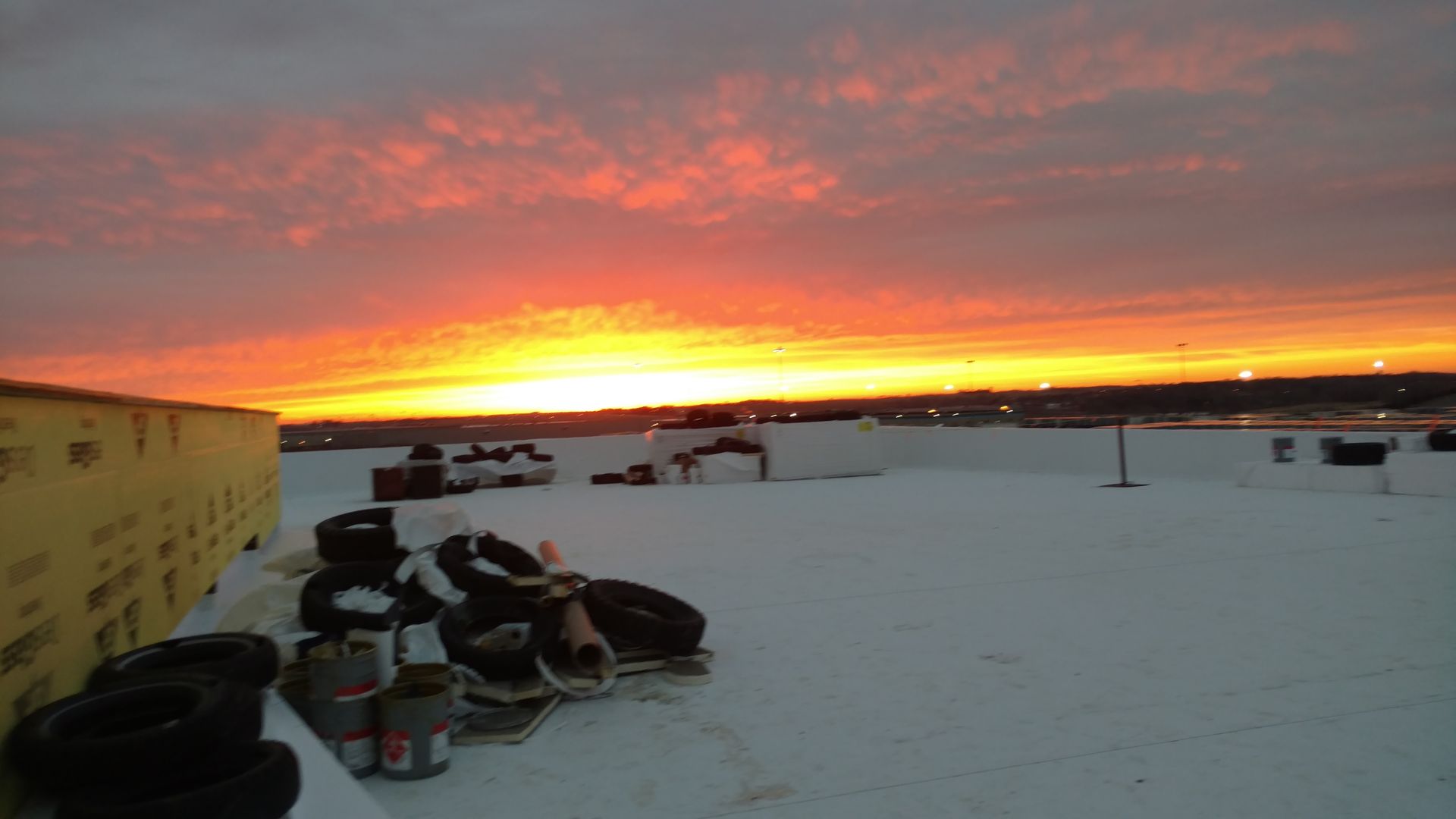 A vibrant, multi-colored sunrise over a snow-covered rooftop with construction materials piled to the side.