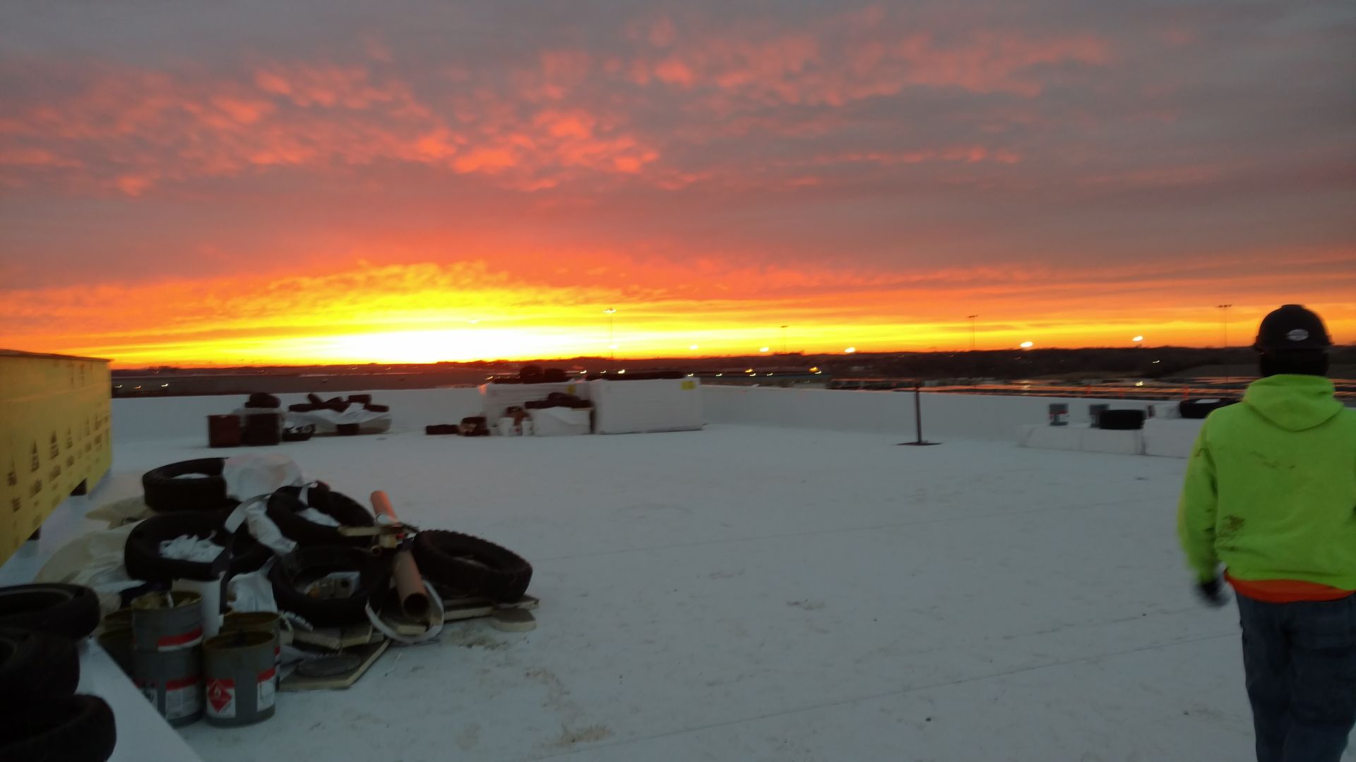 A person in a bright green jacket stands on a white rooftop, looking out at a vibrant orange and yellow sunset.