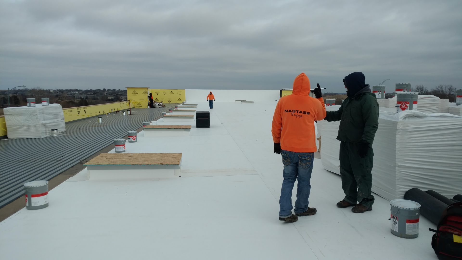 Two workers in high-visibility and dark gear standing on a flat, white rooftop with construction materials nearby.