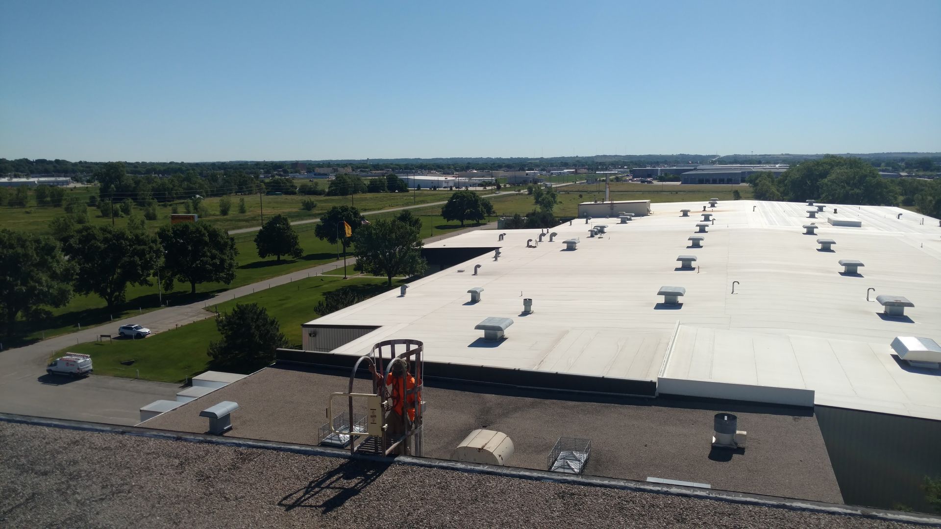 A worker in a safety harness stands on a roof edge, looking out over a large white-roofed building and rural landscape.