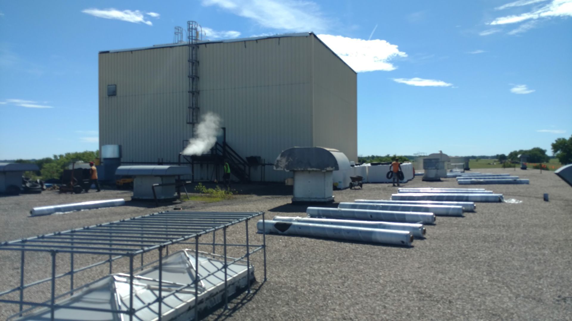A flat, graveled commercial rooftop with metal ventilation ducts and a large beige utility building under a sunny sky.