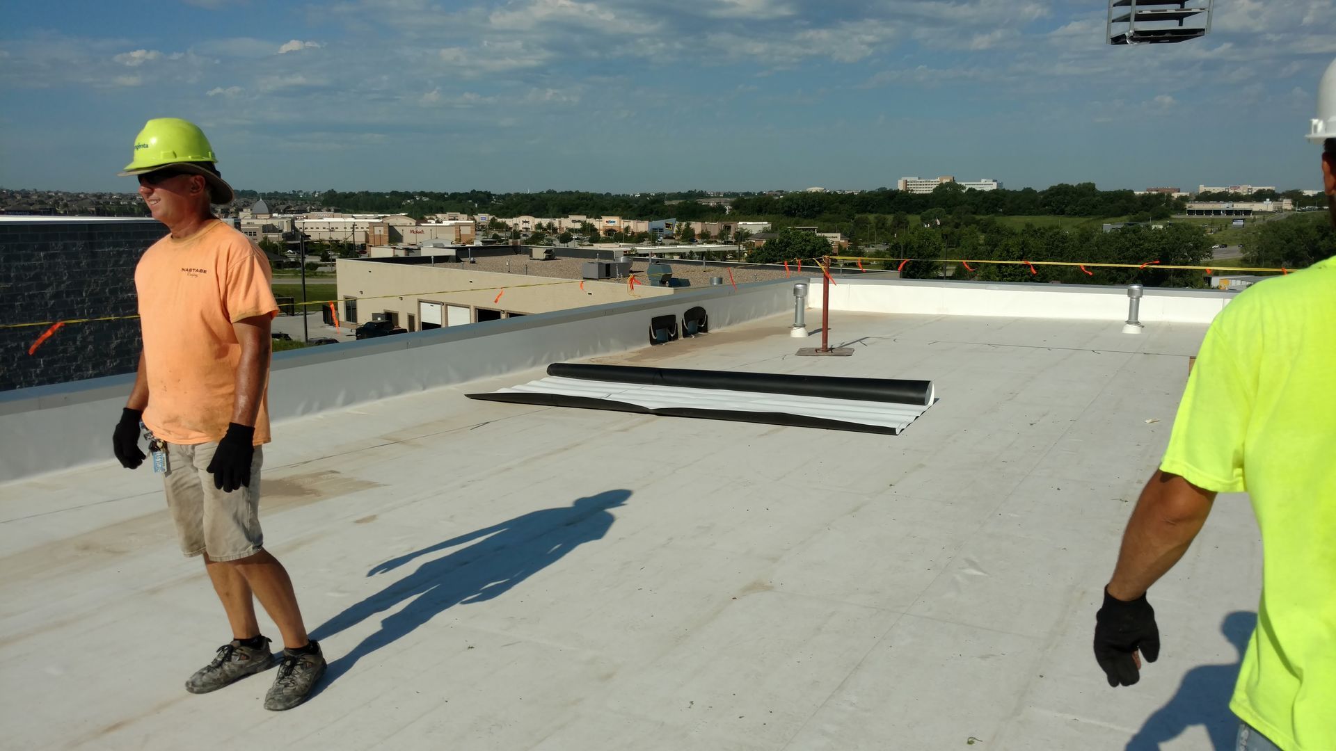Two workers in high-visibility hard hats stand on a flat, white industrial rooftop with a roll of roofing material.