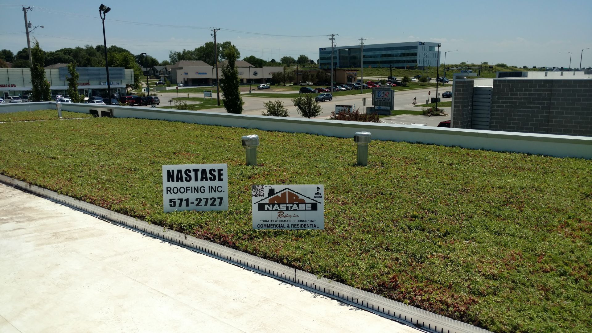 A rooftop covered in green sedum plants featuring two contractor signs in a sunny, suburban commercial setting.