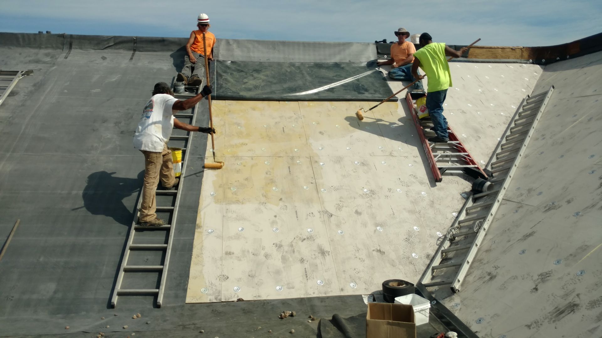 Workers in high-visibility vests install roofing materials on a sloped roof using ladders and tools.