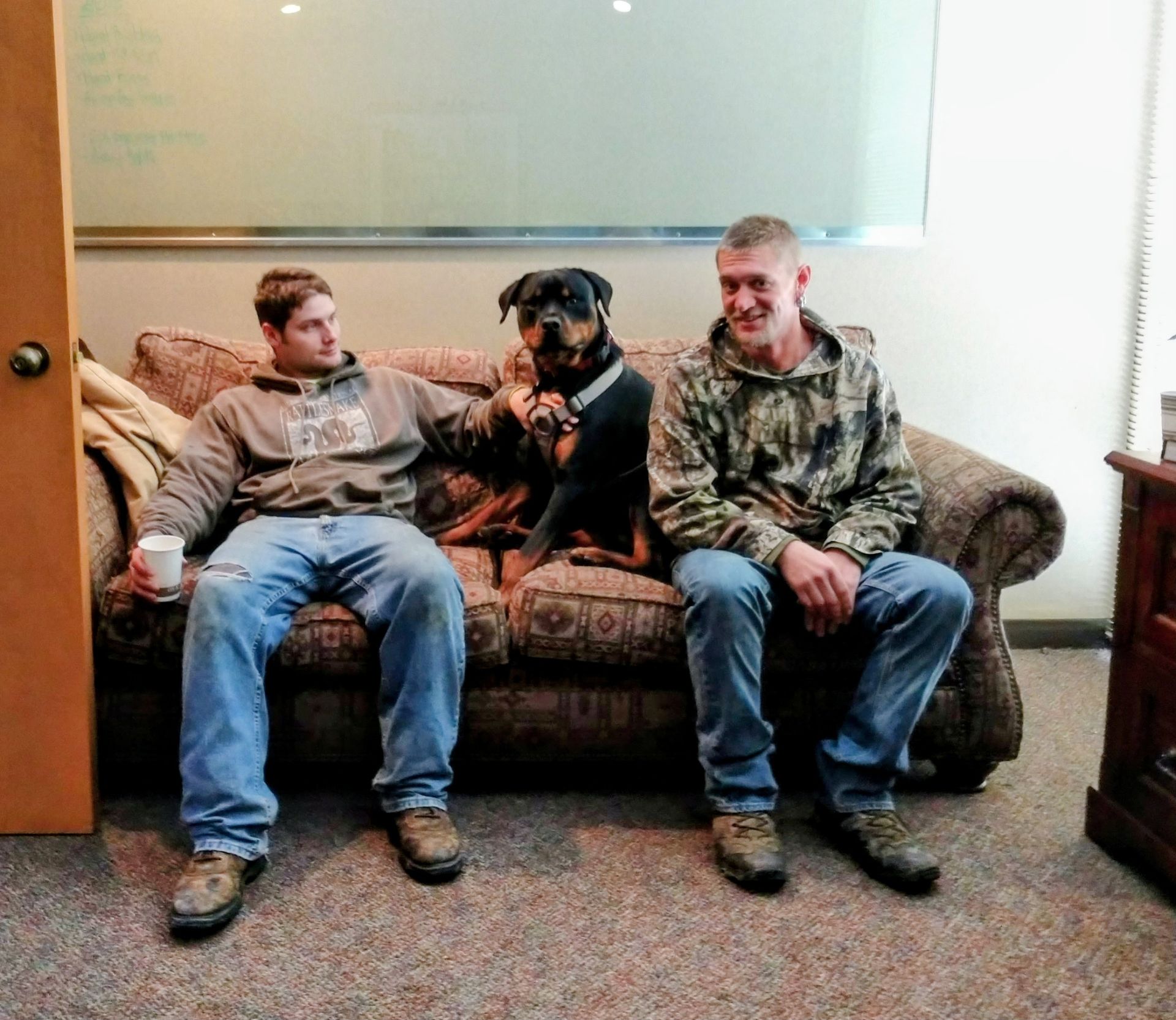 Two people sitting on a patterned couch with a Rottweiler between them in an office setting.