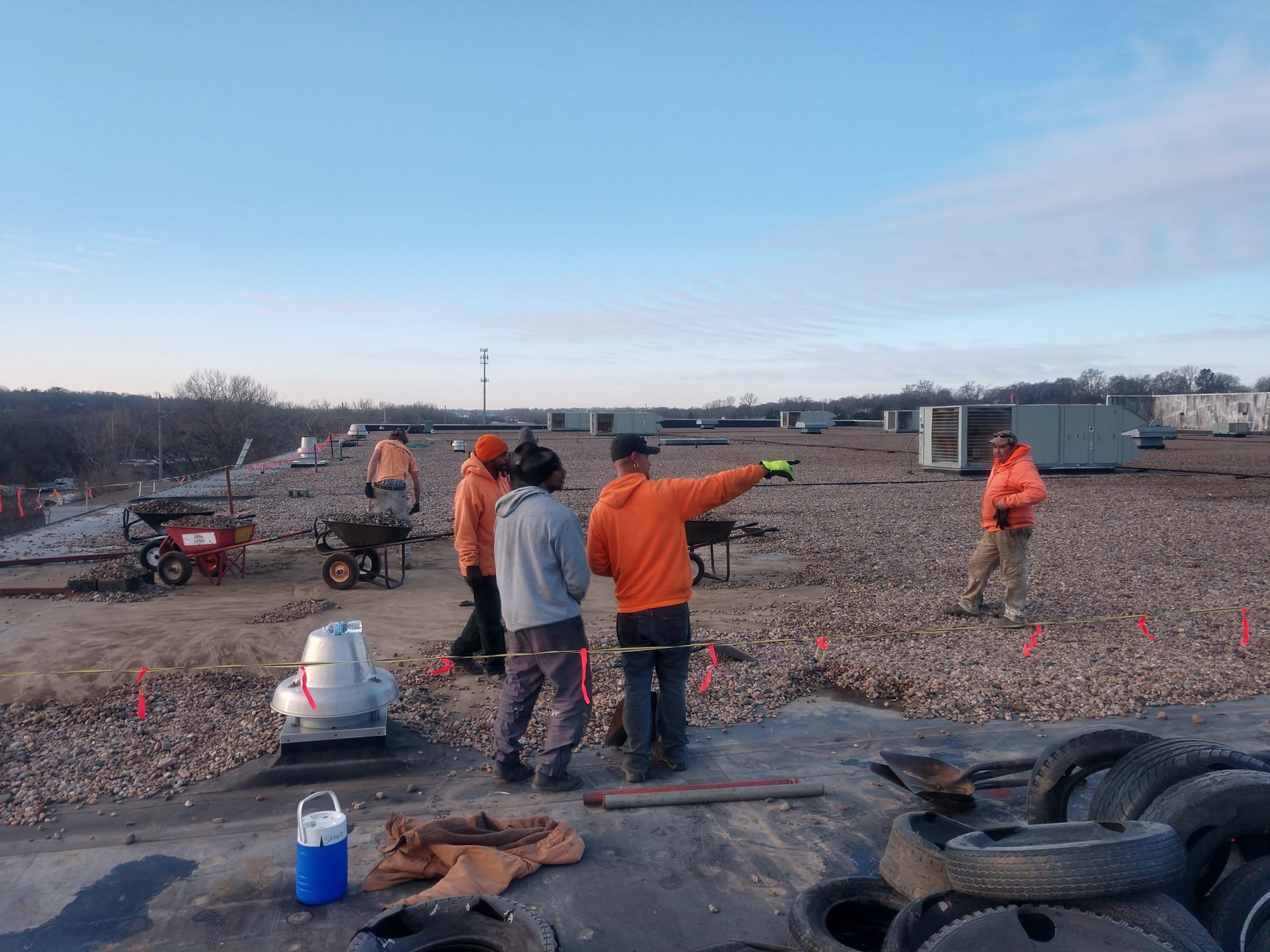 Construction workers in safety gear stand on a flat, gravel-covered roof, one pointing toward HVAC units on a clear day.