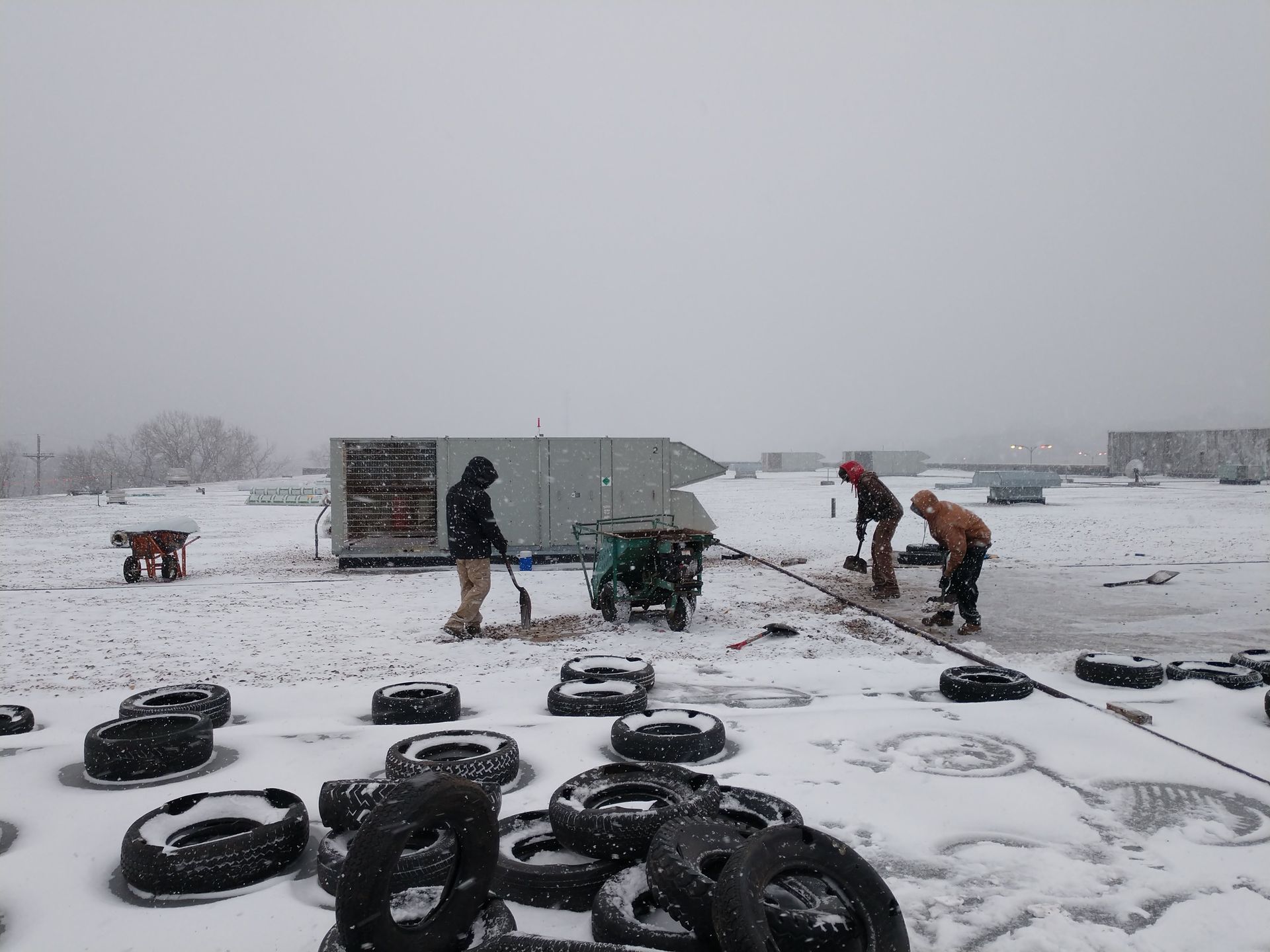 Workers in winter gear shovel snow and use equipment on a snowy rooftop near several discarded tires.