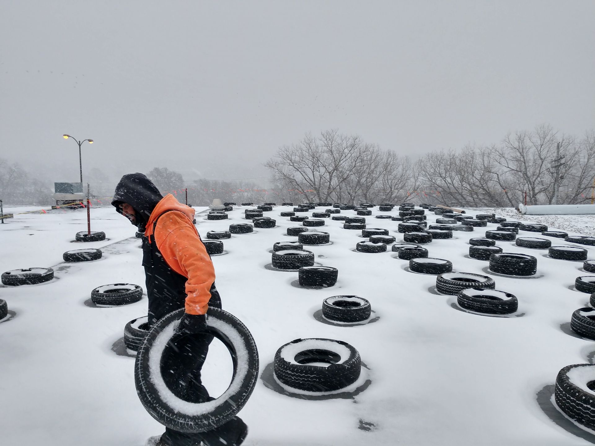 A person in an orange jacket carries a black tire across a snowy lot filled with scattered tires.