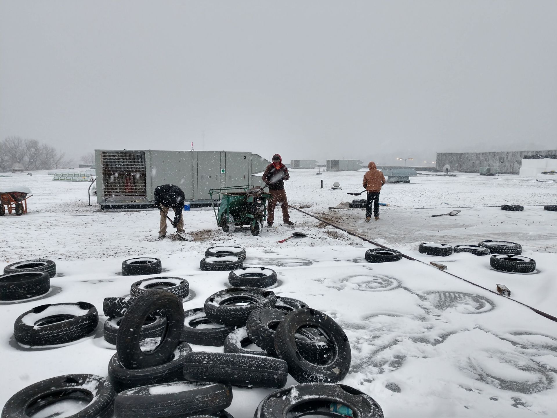 Workers clear snow near several scattered tires in a snowy, industrial outdoor lot.
