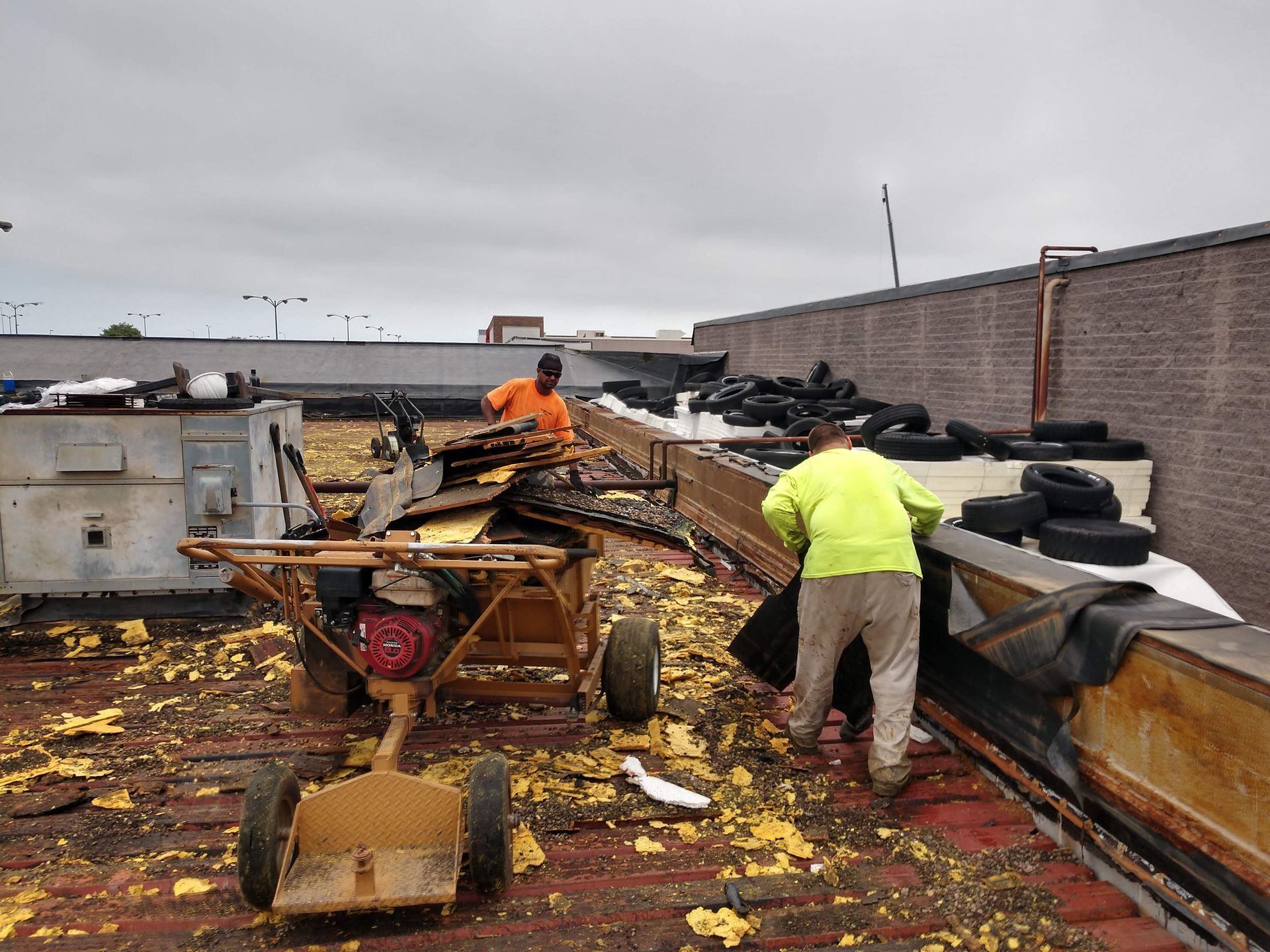 Two workers in high-visibility shirts tear up old roofing materials on a commercial building roof.