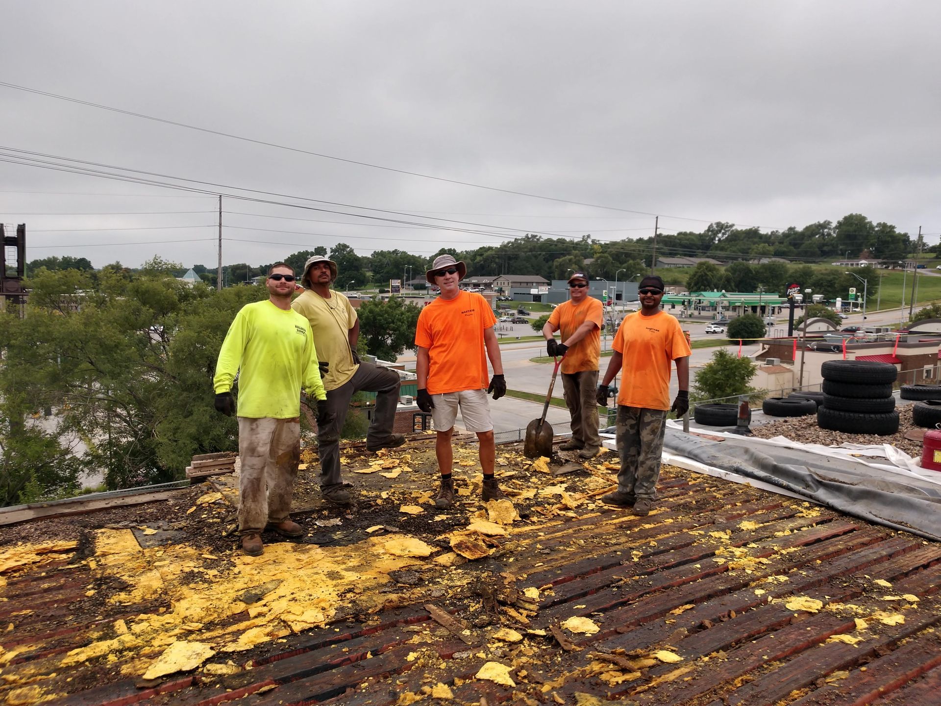 Five workers in safety gear stand on a commercial rooftop with yellow debris, posing for a photo under an overcast sky.