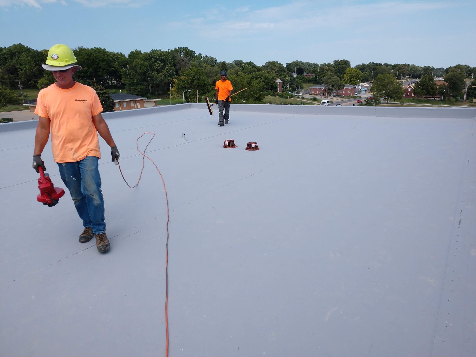 Two workers in high-visibility shirts inspect a flat roof surface using specialized handheld equipment.