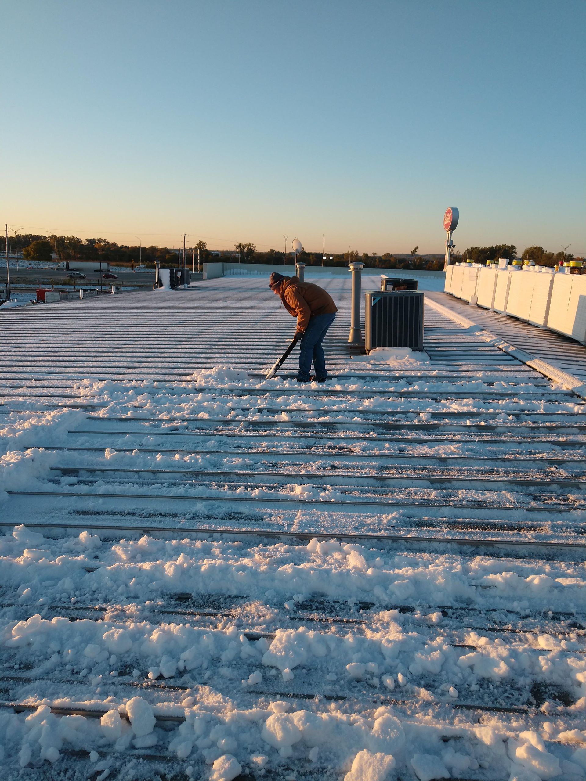 A person in a brown jacket shovels snow off a wooden pier under a bright, clear blue sky at sunset.