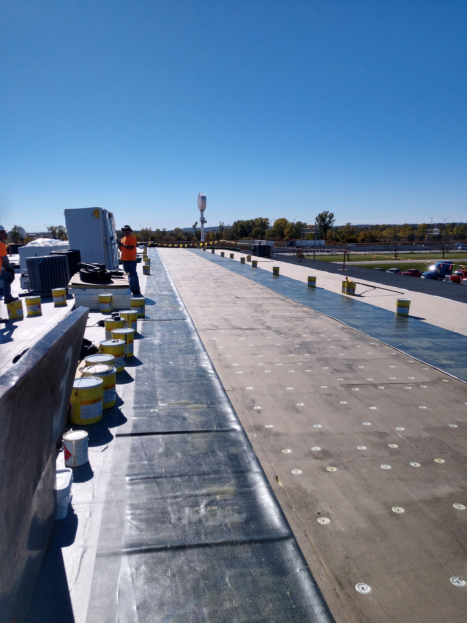 Workers on a commercial roof applying dark, flat roofing membrane over a surface with grid-patterned white adhesive spots.