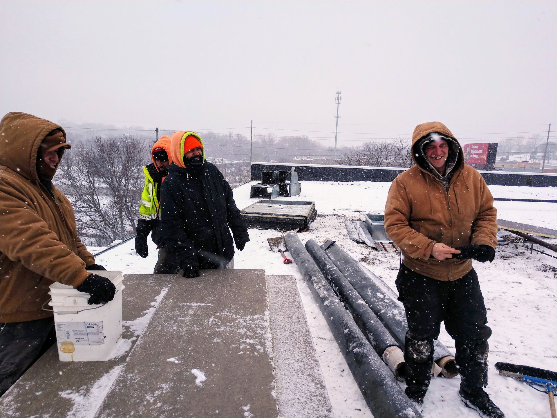 Construction workers wearing heavy winter gear and hoods on a snow-covered rooftop while working on materials.