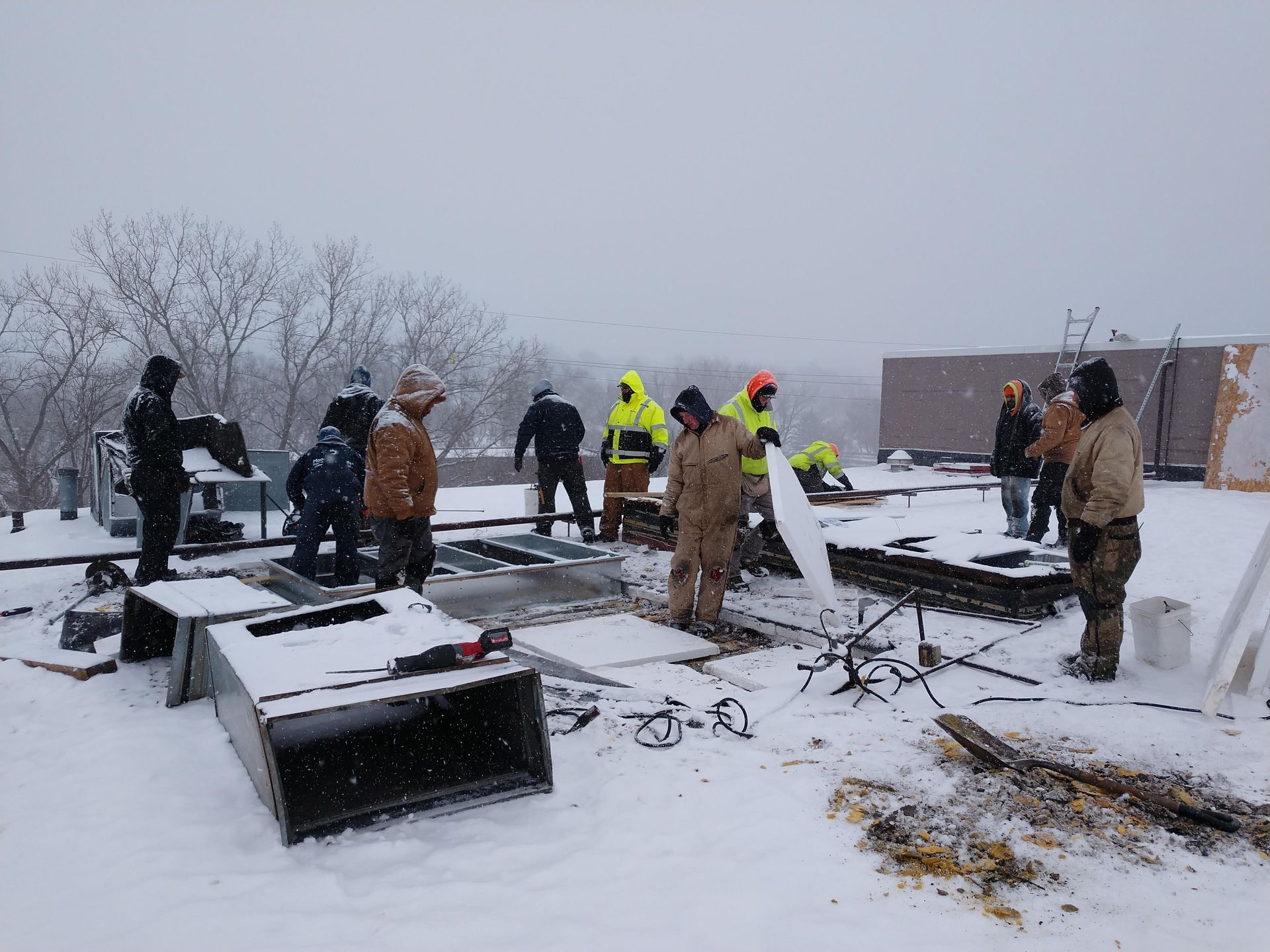 A group of workers in heavy winter gear perform construction work on a snow-covered flat rooftop.