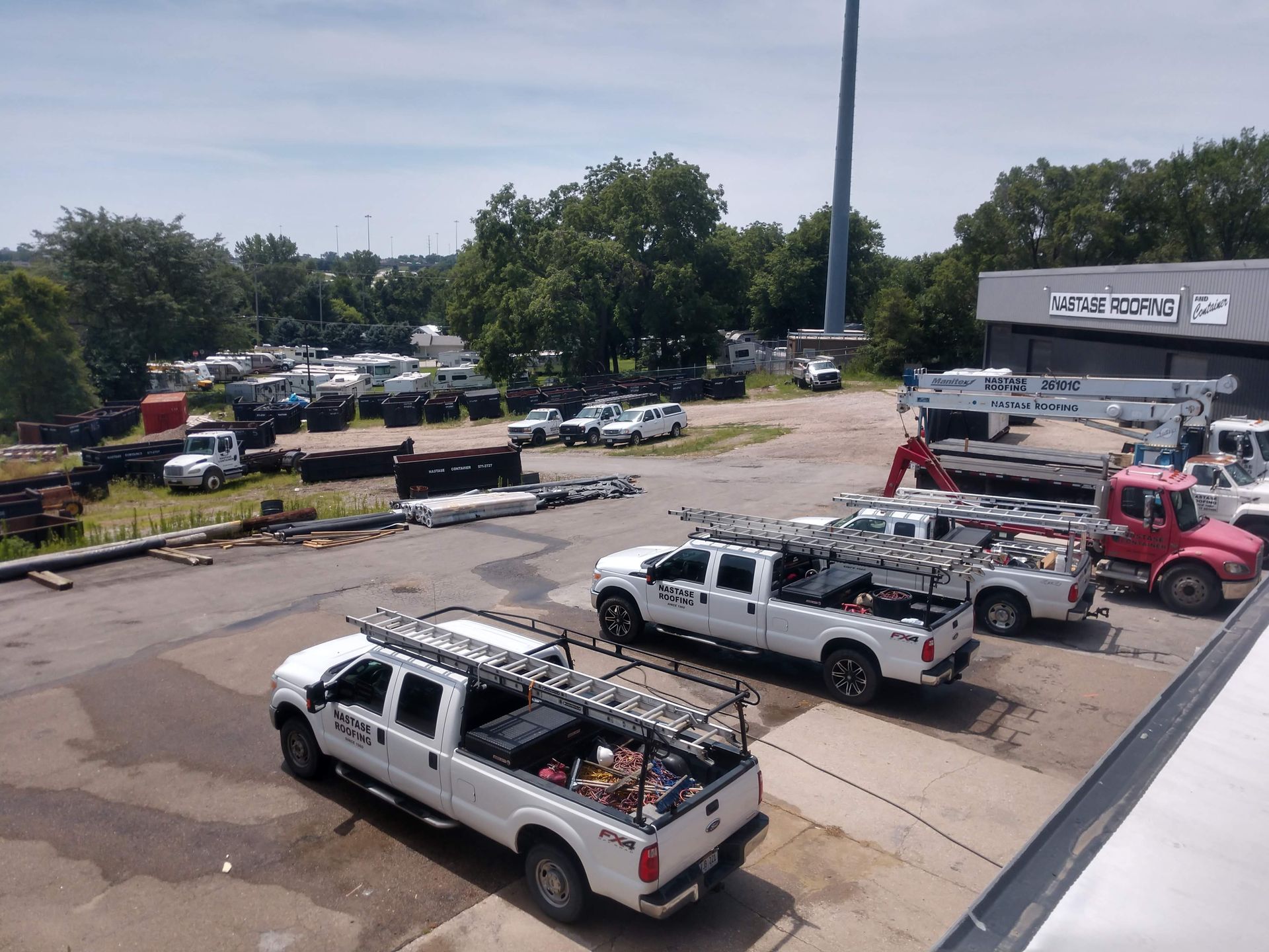 A fleet of white work trucks with ladder racks parked in a lot beside a building, with a crane truck in the background.