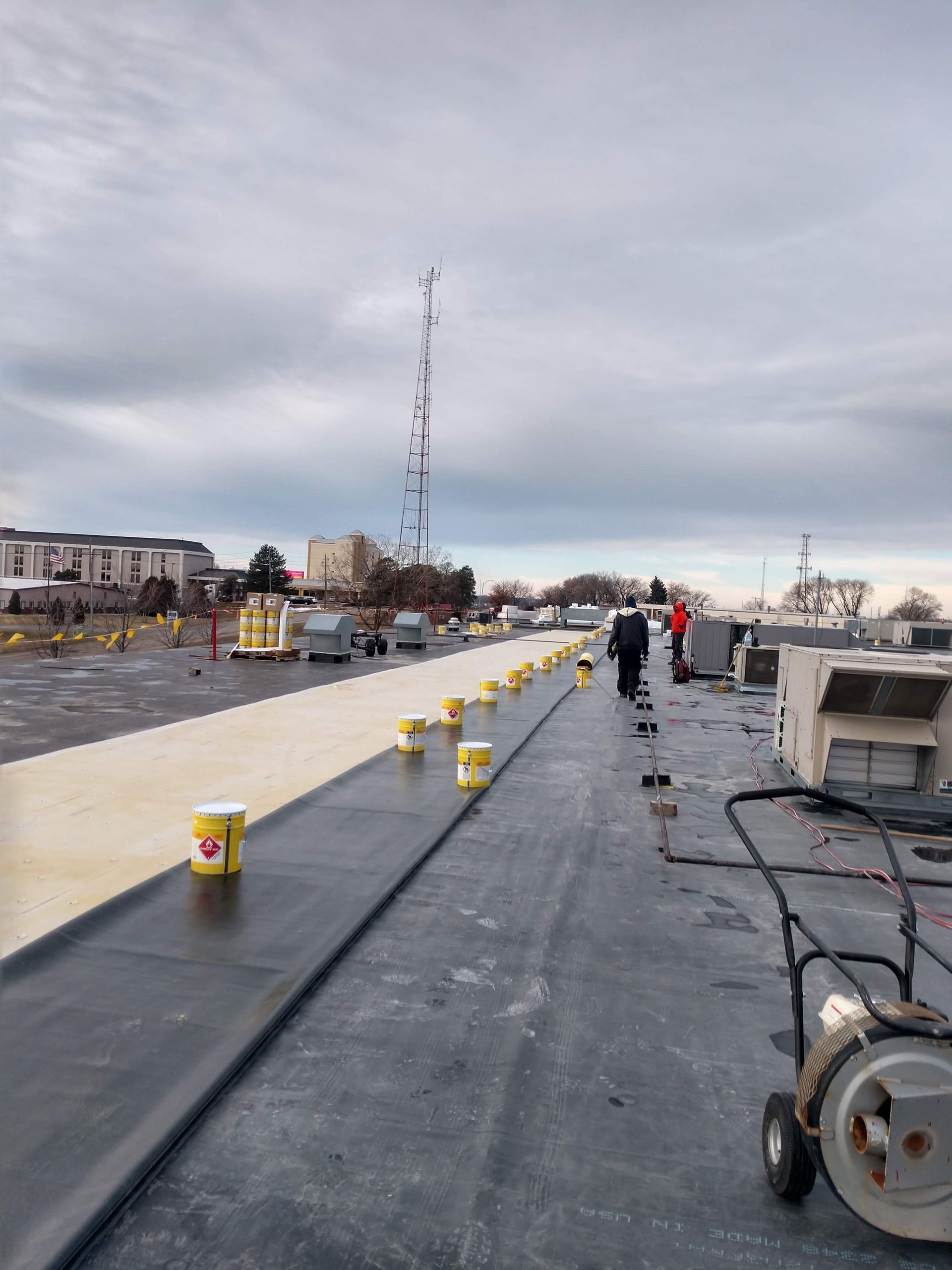 Workers apply sealant to a flat roof, featuring a row of yellow buckets on a dark surface under a cloudy sky.