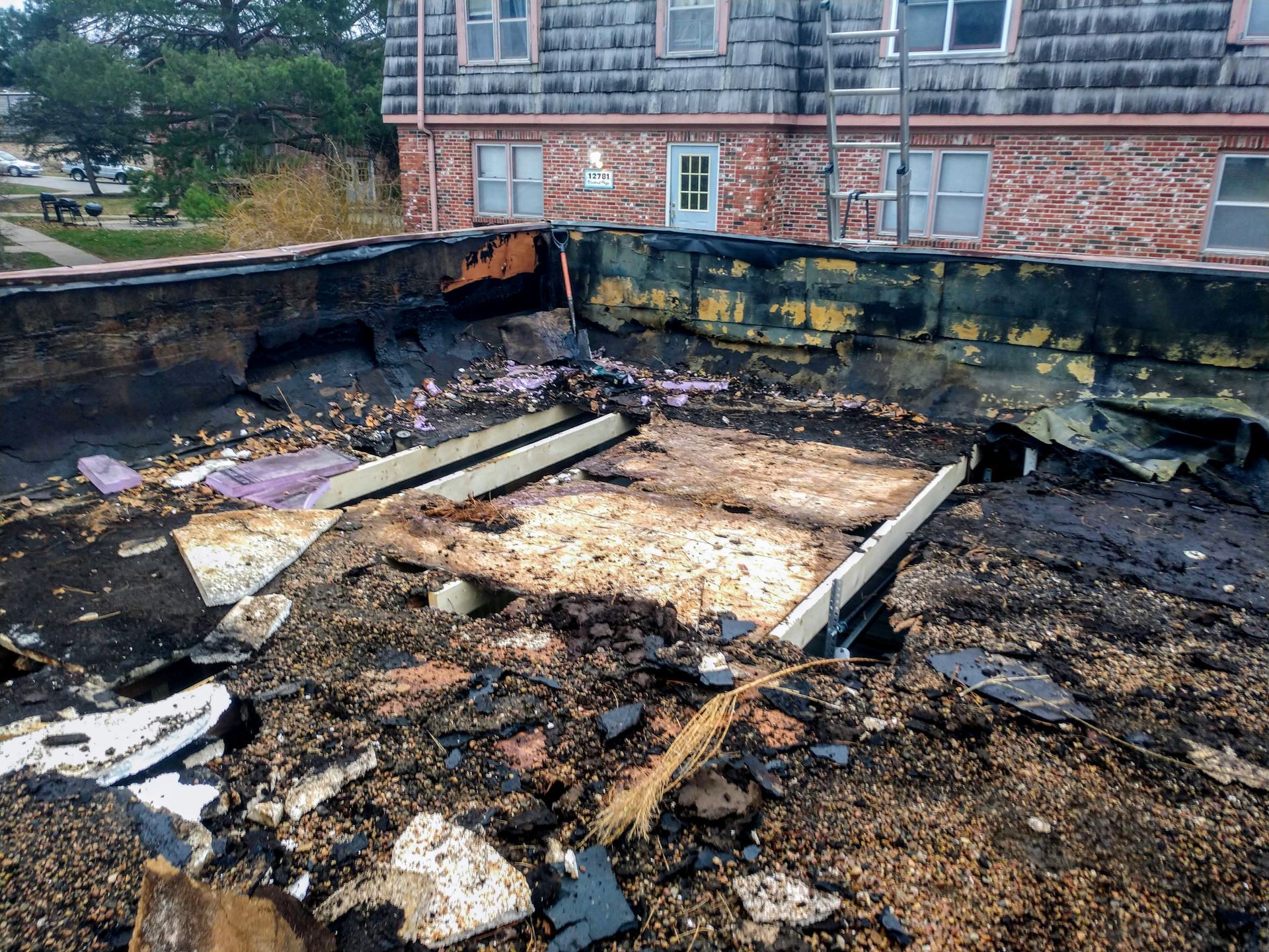 A roof undergoing demolition with exposed wooden decking and scattered debris, set against the side of a brick building.