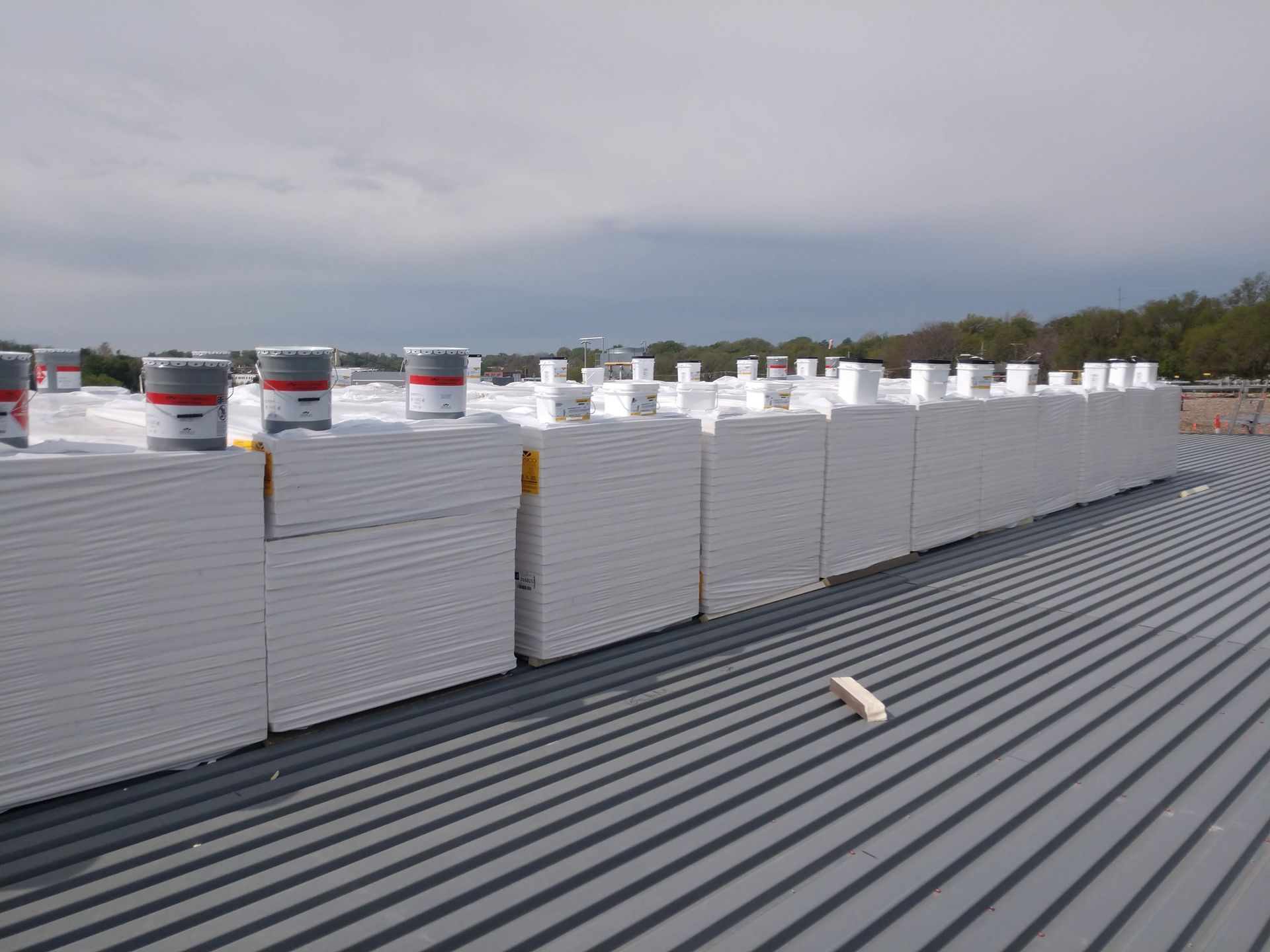 Stacks of drywall boards and buckets of joint compound sit on a metal roof under a cloudy sky.