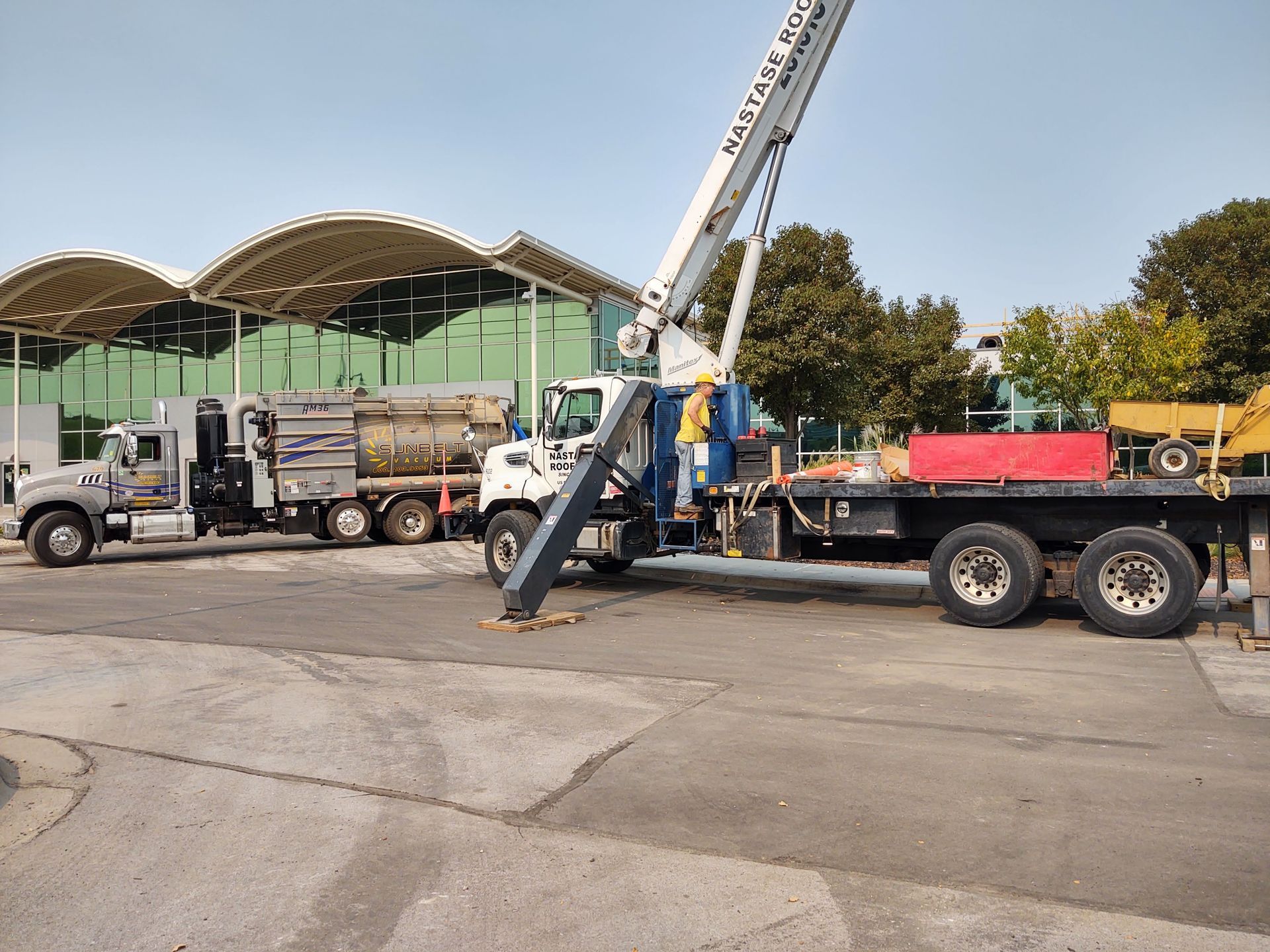 A crane truck parked on a paved lot with a worker near the crane base, next to a trailer carrying industrial equipment.