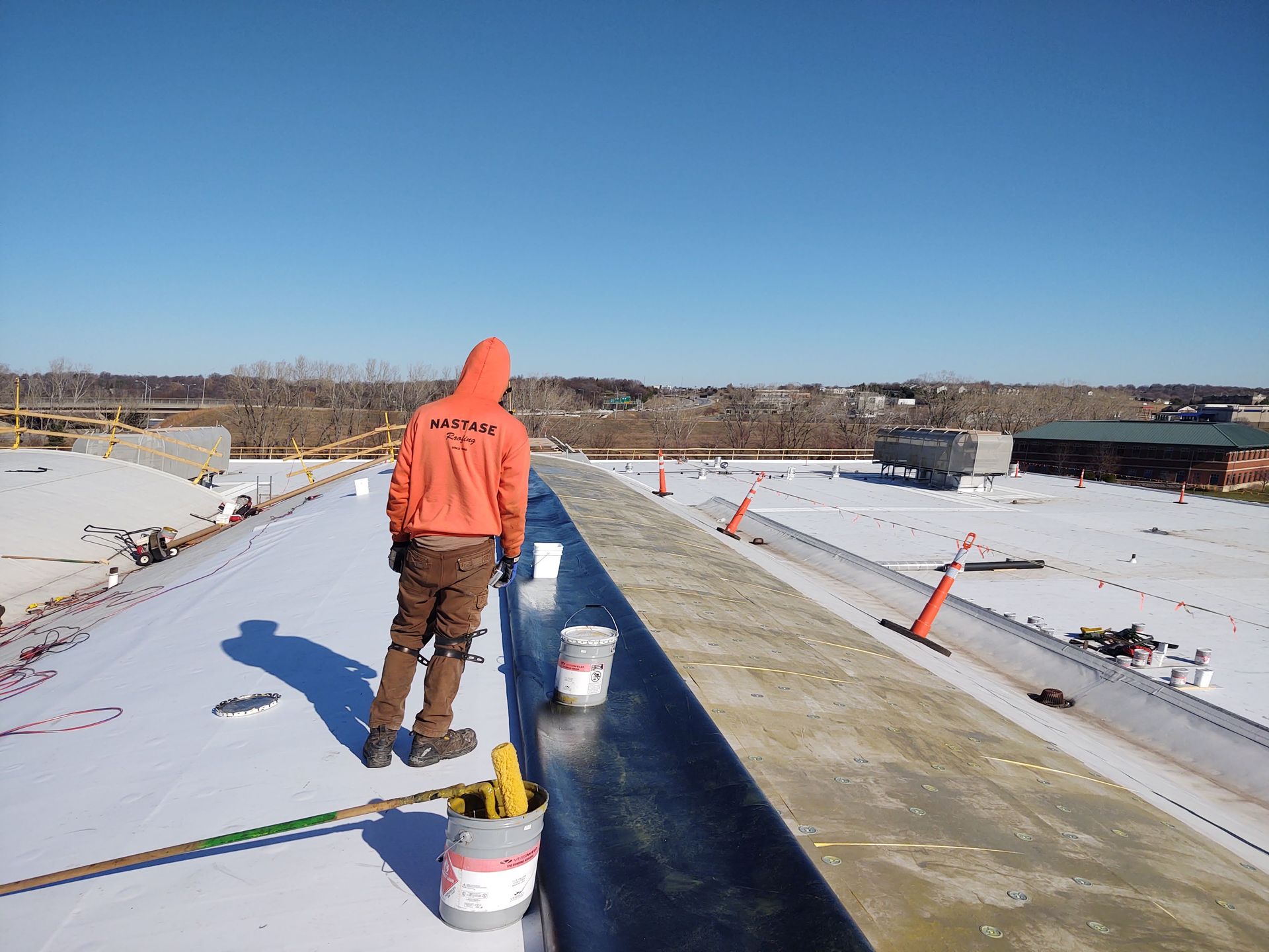 A worker in an orange hoodie installs dark roofing material on a sunny, white rooftop construction site.