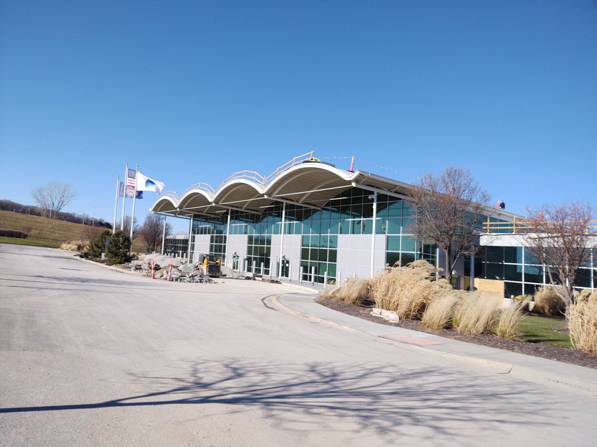 The modern, glass-walled building features a wavy, white roof, set beside a paved road under a clear blue sky.