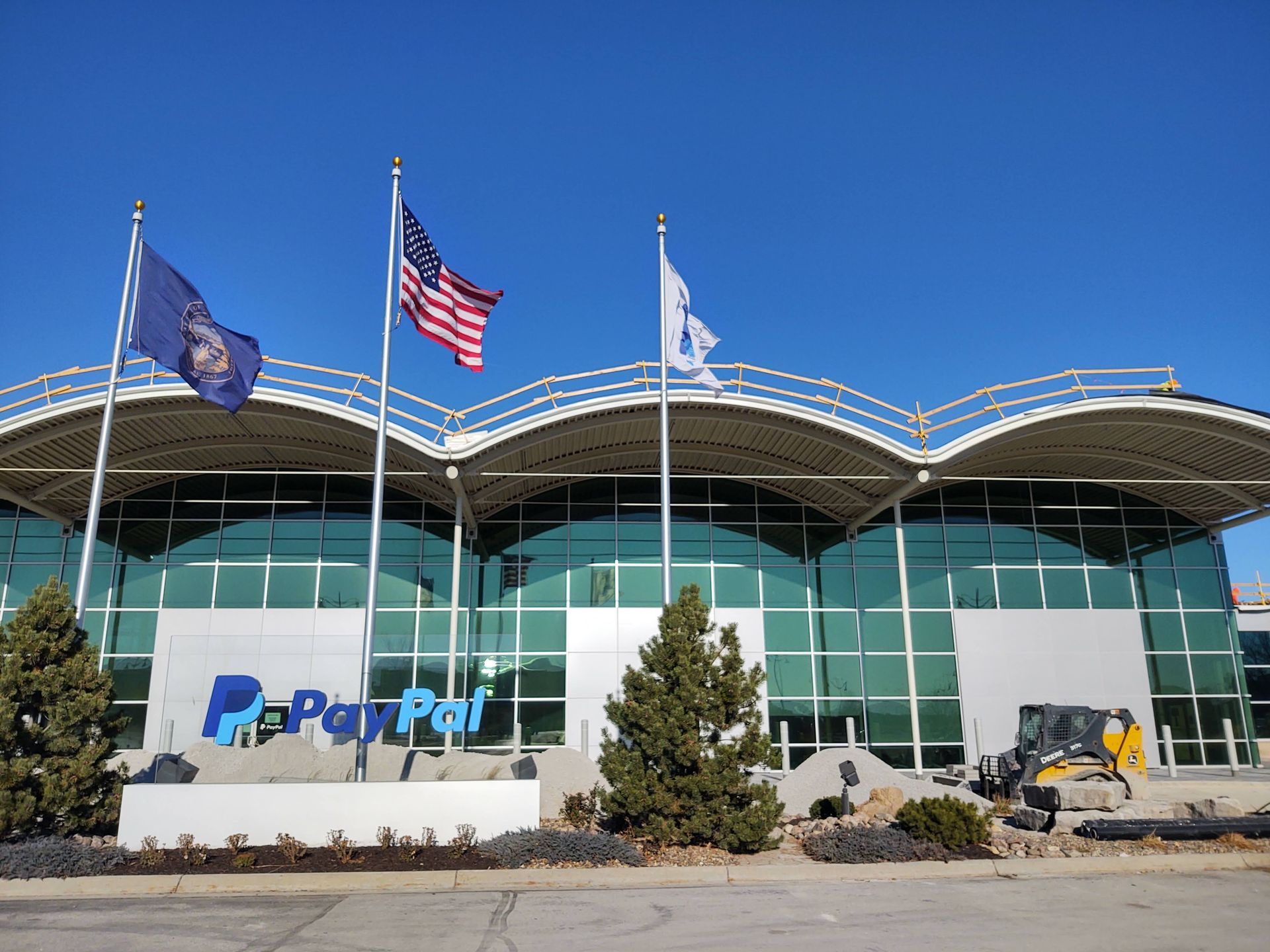 PayPal office building with a wavy roofline, three flags, and a construction vehicle in front under a clear blue sky.