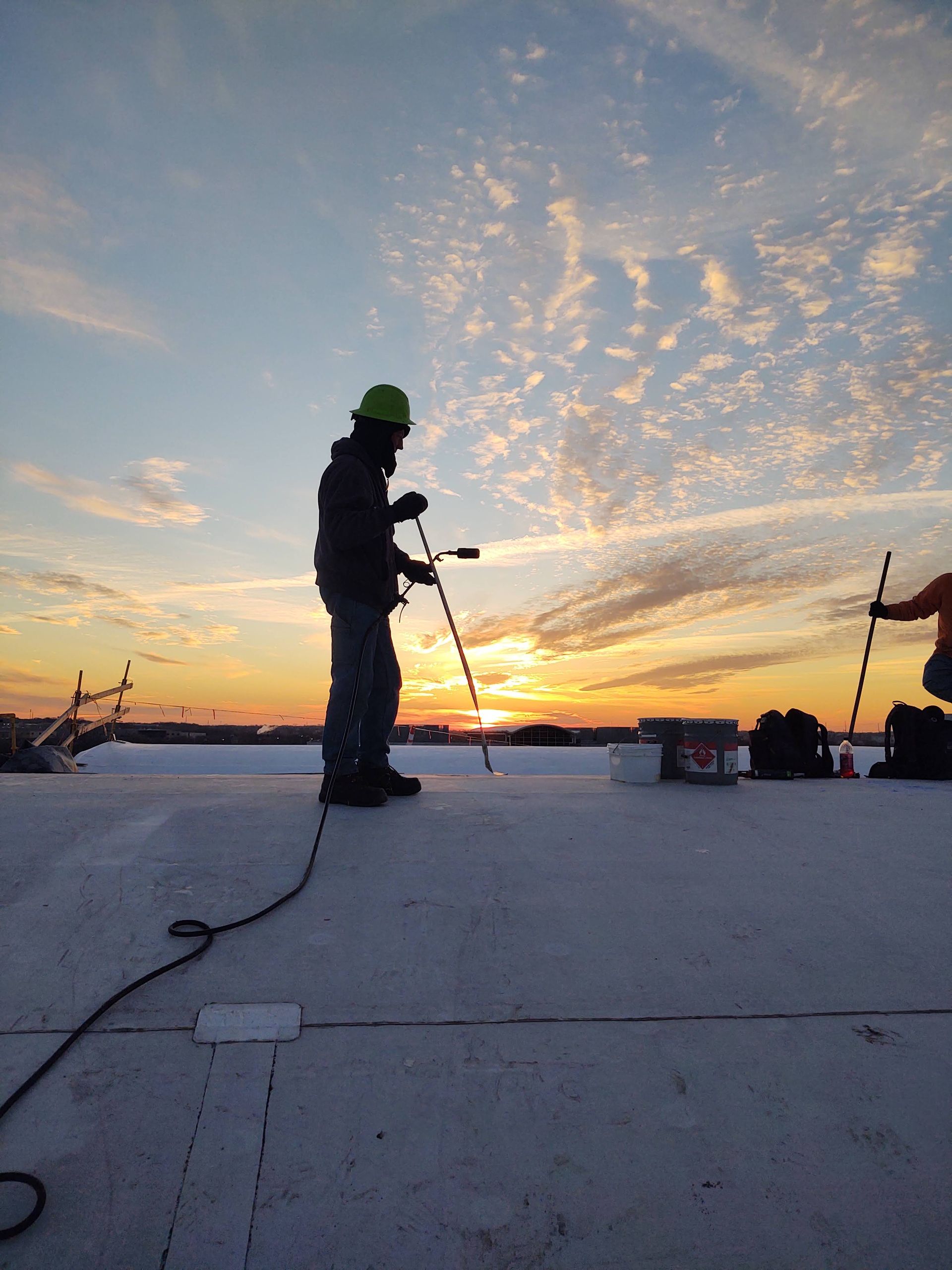 Silhouetted construction workers on a flat roof at sunset, using tools to install roofing materials.