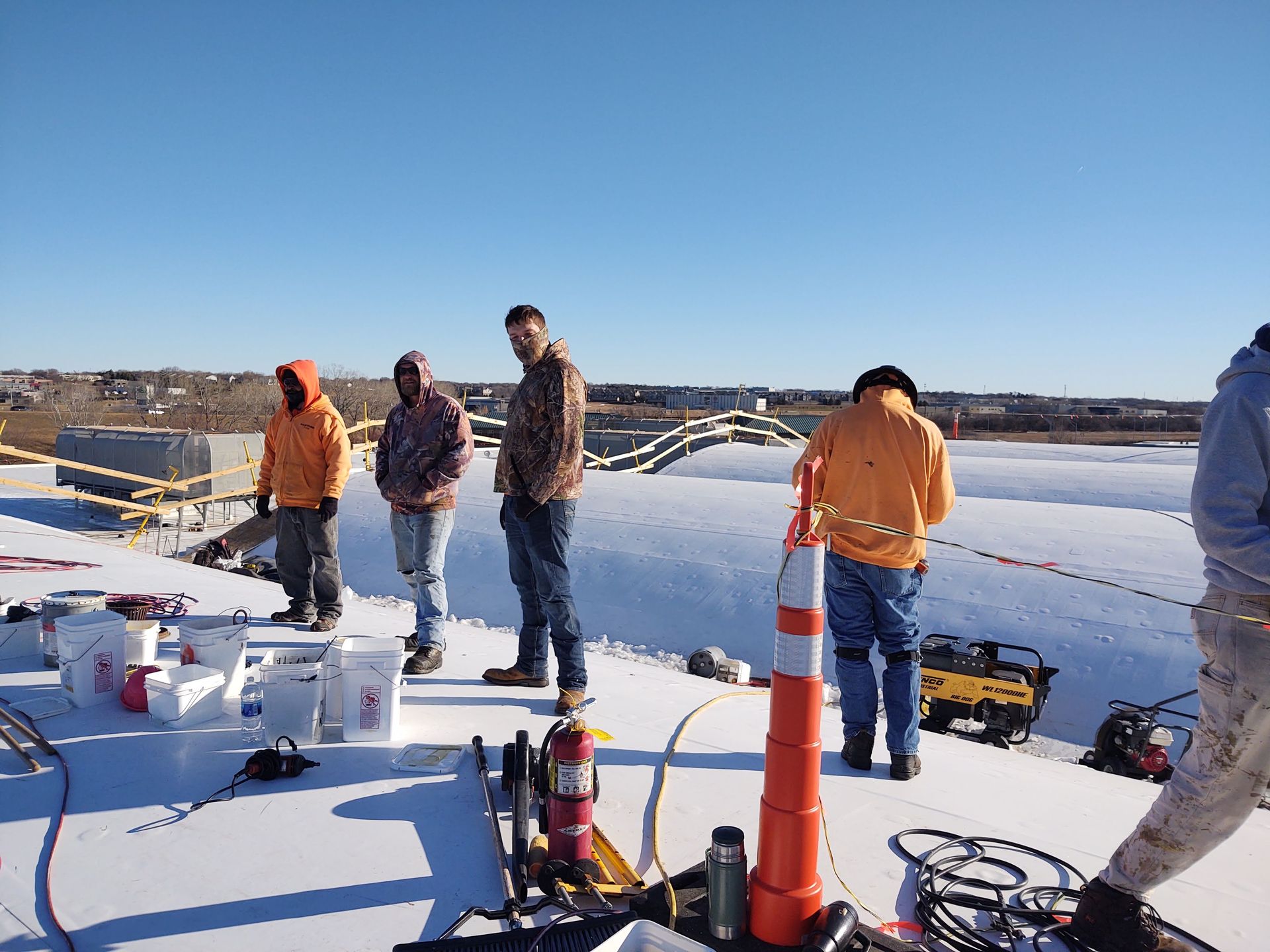 Construction workers in winter gear stand on a white snow-covered roof near tools, equipment, and an orange traffic cone.