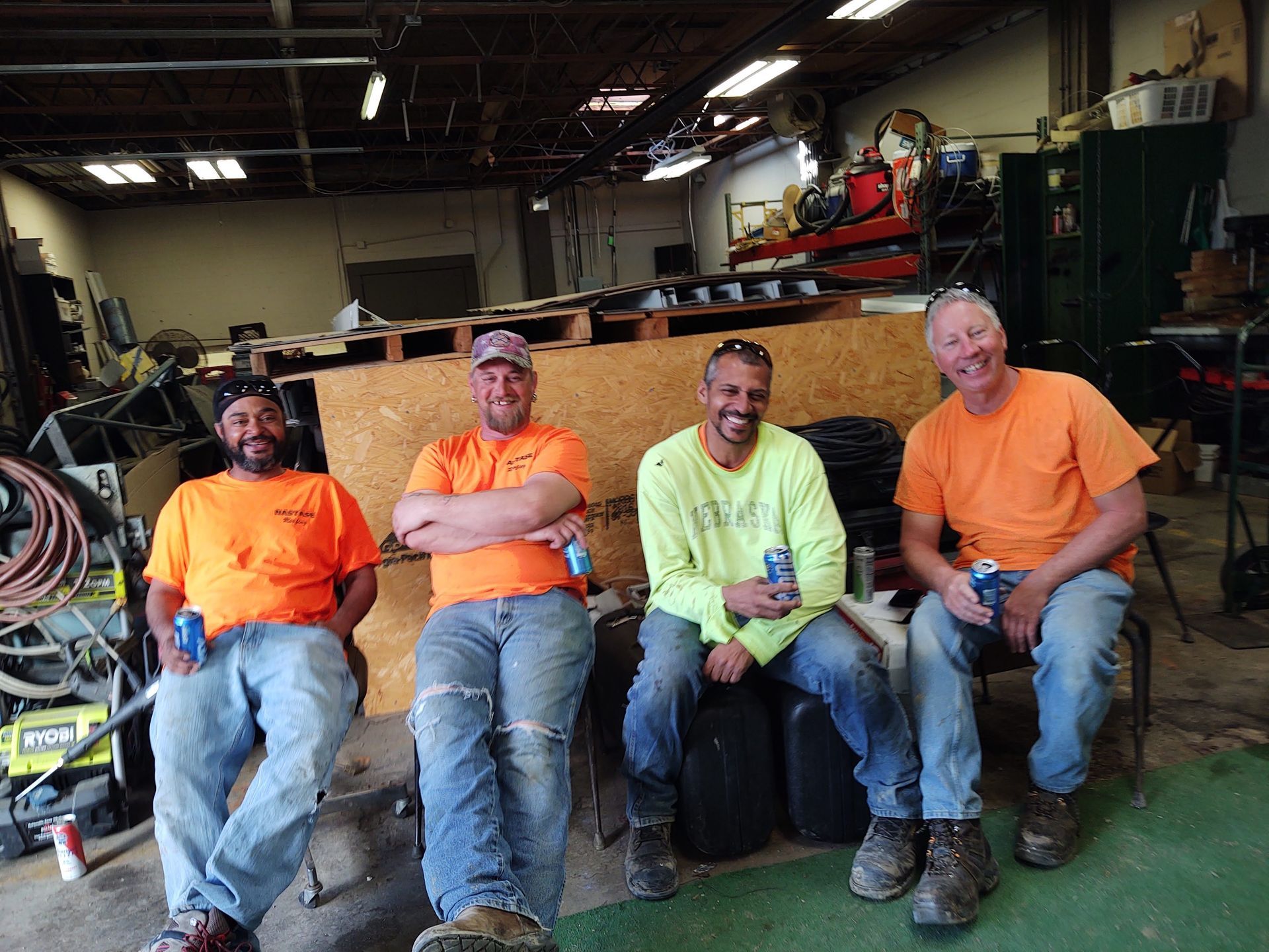Four people wearing work clothes sit in a garage, smiling and holding drinks.