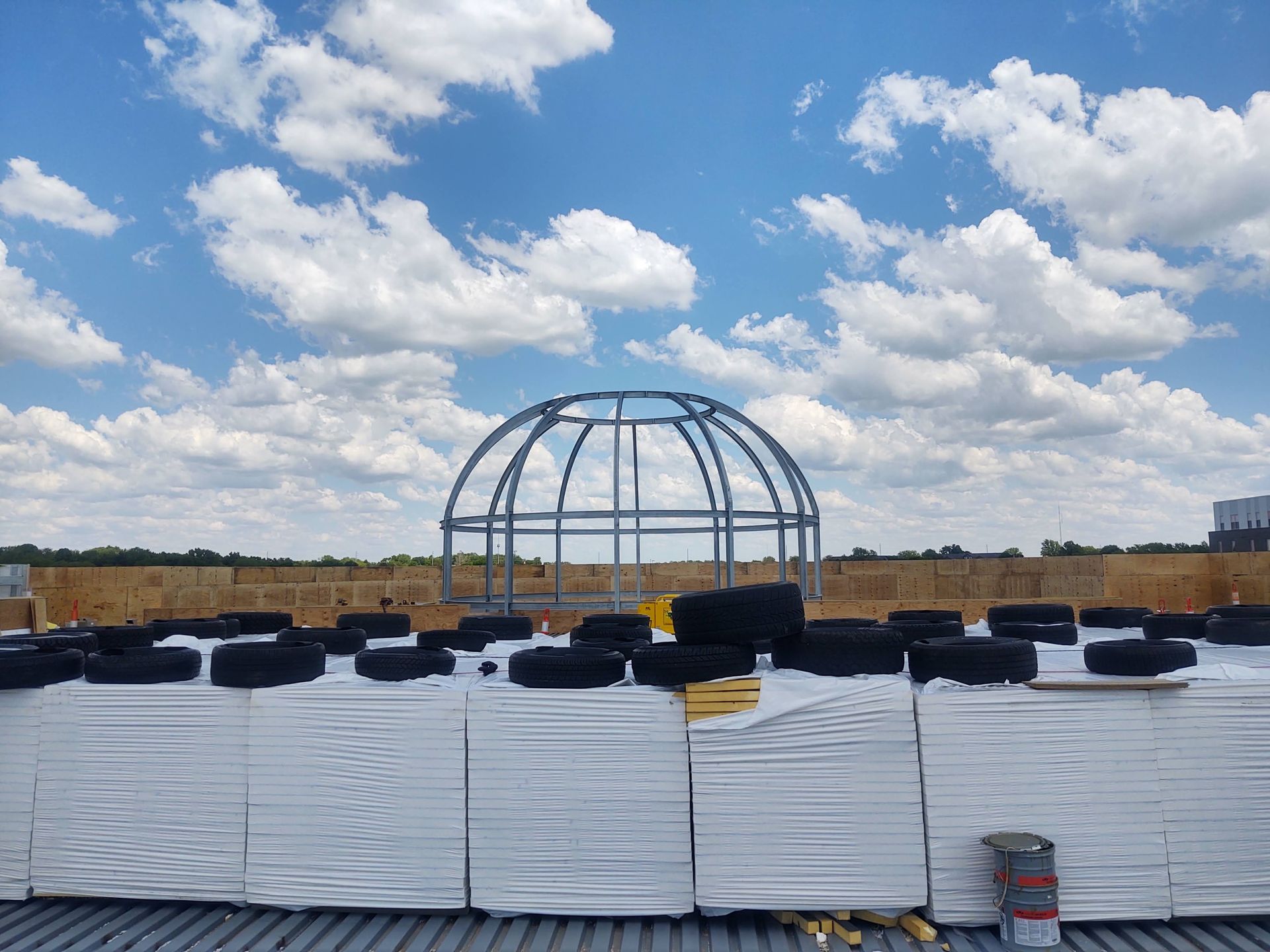 A metal dome frame stands on a rooftop behind stacked construction materials under a blue, cloudy sky.
