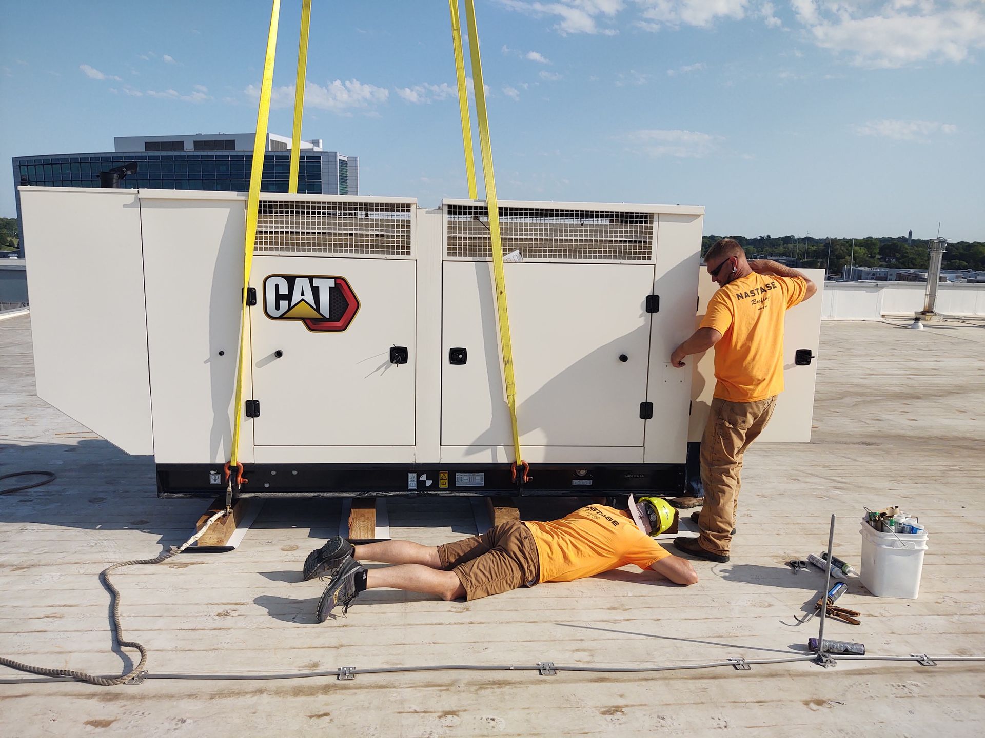 Two workers in yellow shirts install a white CAT generator suspended by yellow crane straps on a rooftop.