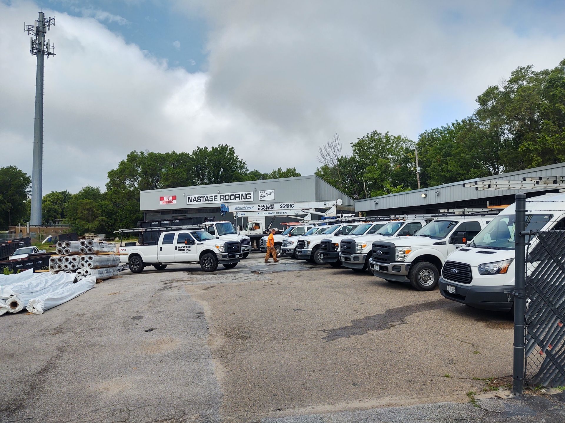 A row of white utility trucks parked in a gravel lot in front of a service building under a partly cloudy sky.