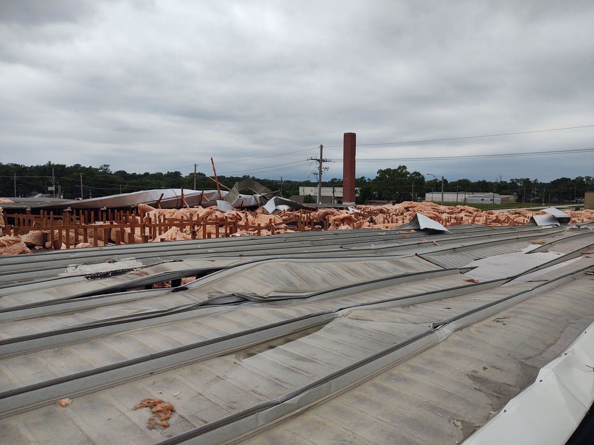 A damaged building rooftop with exposed structural beams, insulation, and debris under a cloudy sky.