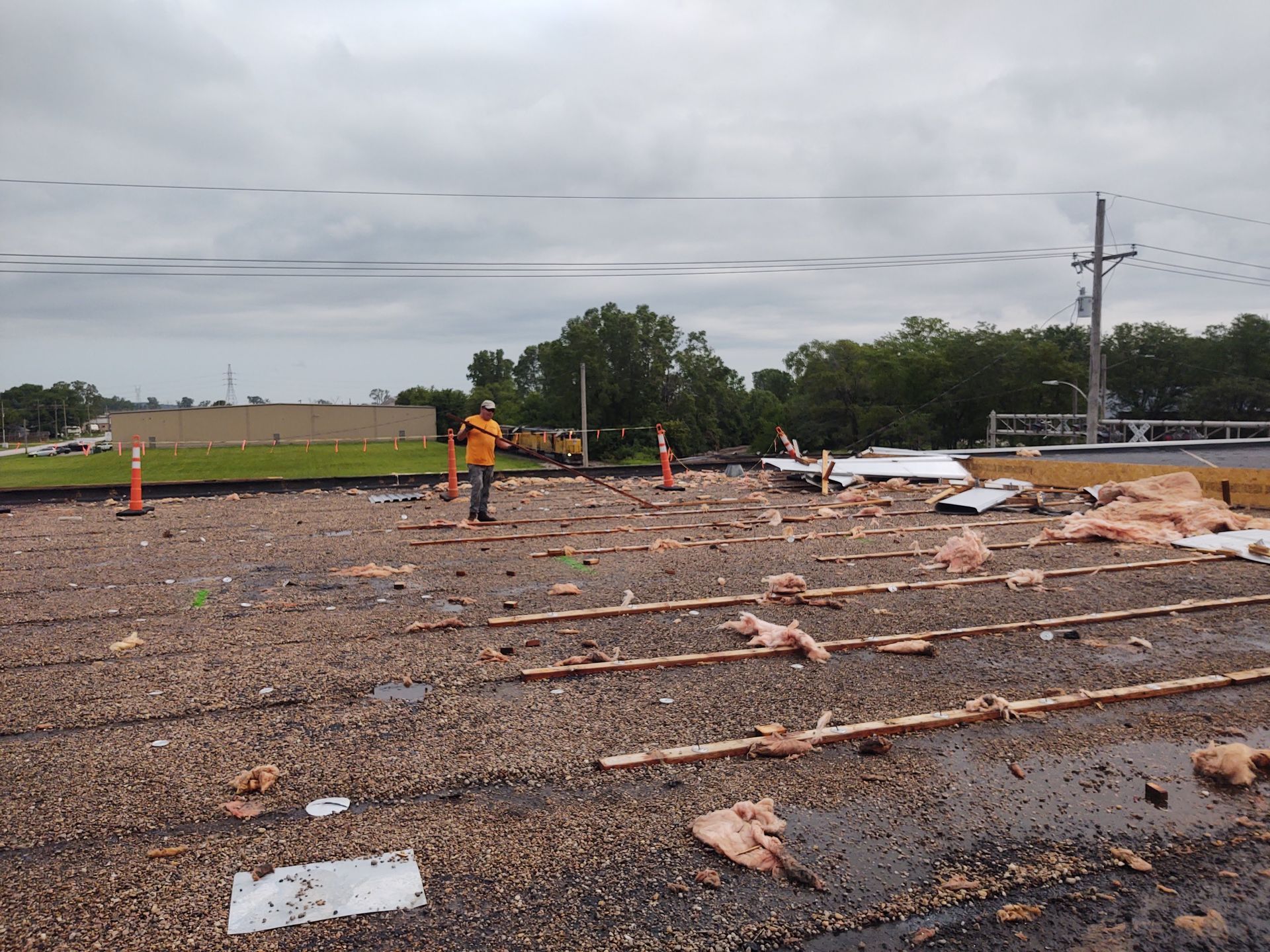 A worker in a yellow shirt stands on a damaged, debris-strewn flat roof under a cloudy sky.