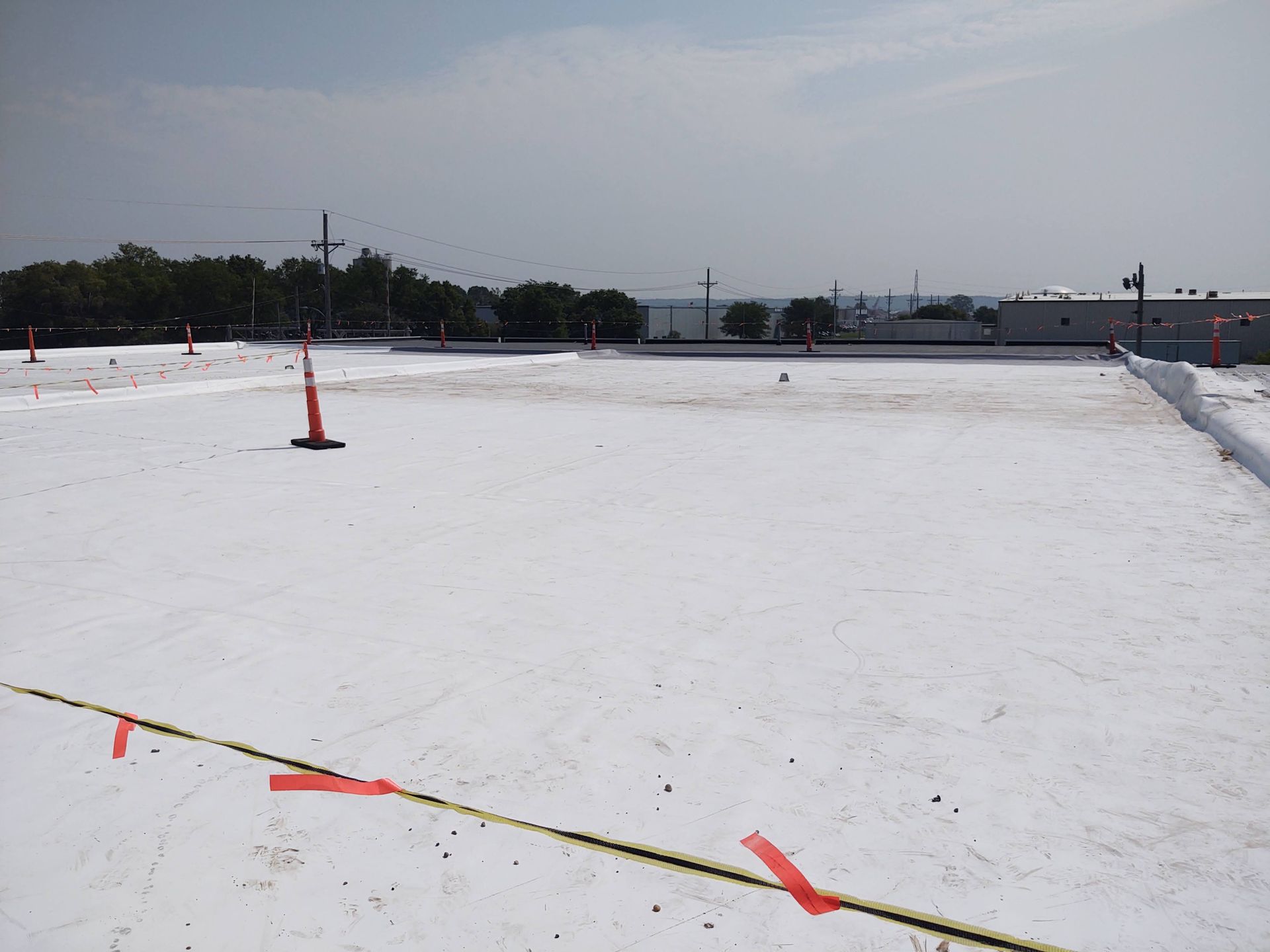 A flat roof with a white protective coating, marked with safety cones and yellow caution tape under a cloudy sky.