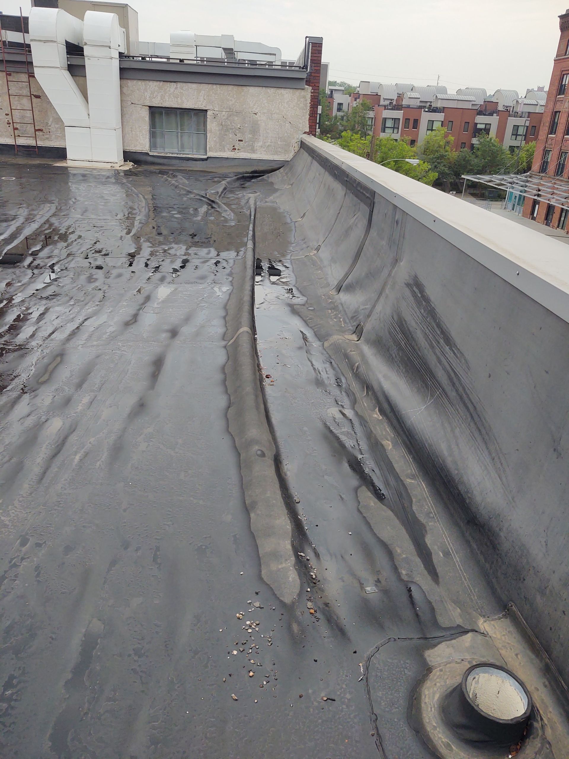 A weathered flat roof with bubbling, black membrane material near a parapet wall, building vents, and distant city views.
