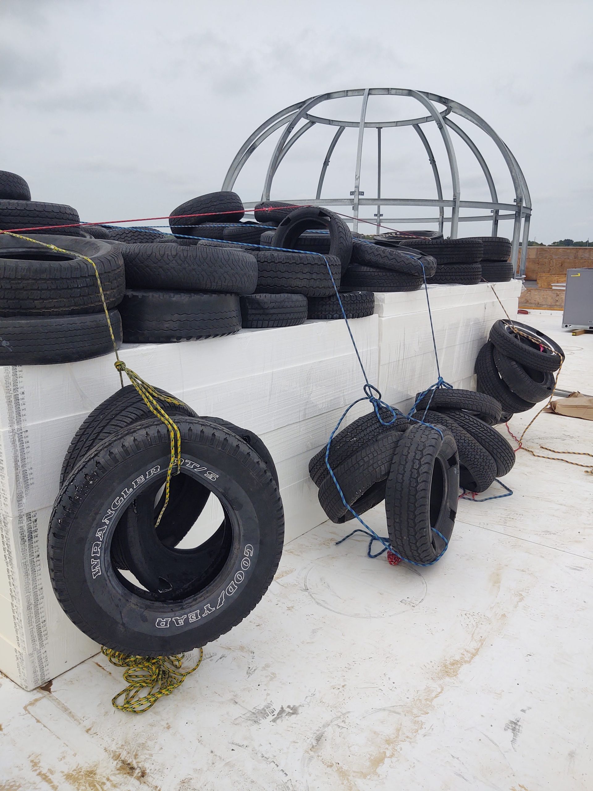 Old tires tied with ropes hanging from a white concrete wall, with a metal dome structure visible in the background.
