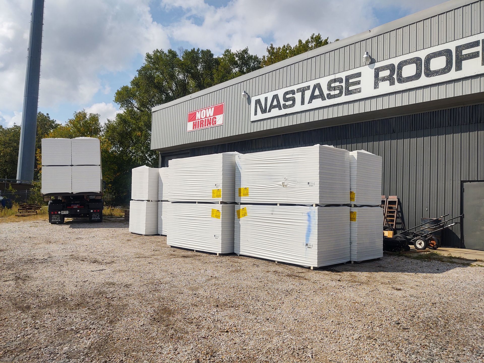 A white trailer sits on a gravel lot next to stacked, plastic-wrapped building supplies outside Nastase Roofing.