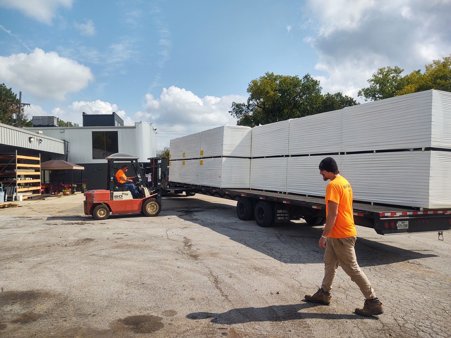 A forklift unloads large, white-wrapped pallet stacks from a flatbed trailer at an industrial yard under a sunny sky.