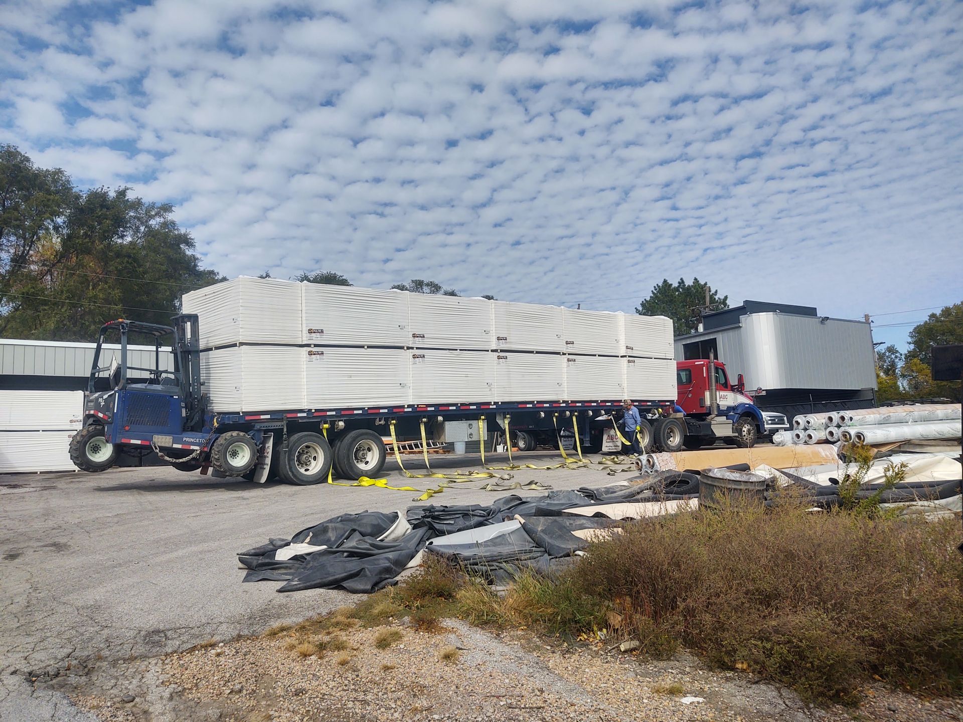 A red semi-truck hauls a flatbed trailer loaded with large white building materials in an outdoor, gravel-filled lot.