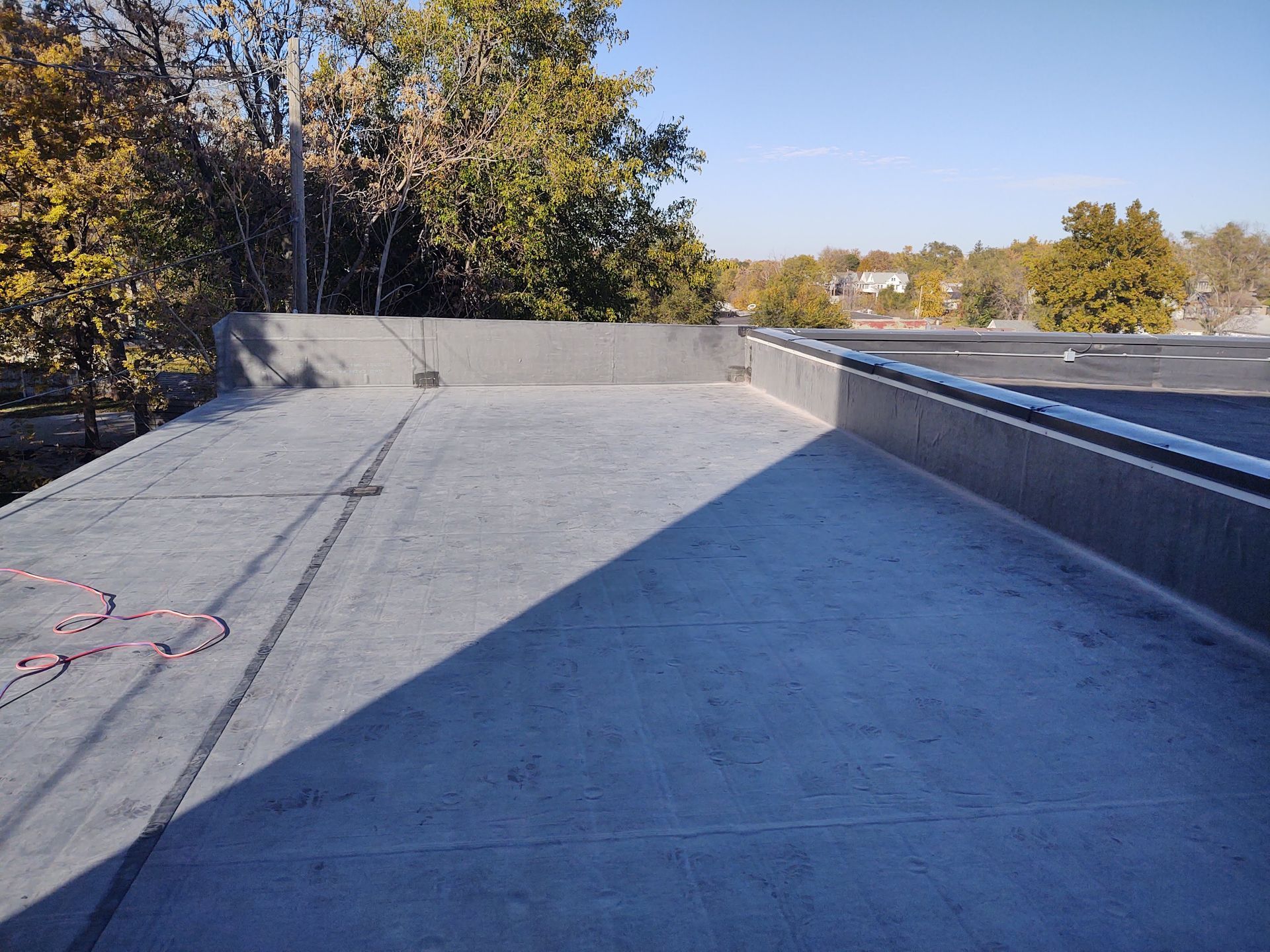 A flat, grey roof surface with a surrounding wall against a backdrop of trees and a clear blue sky.