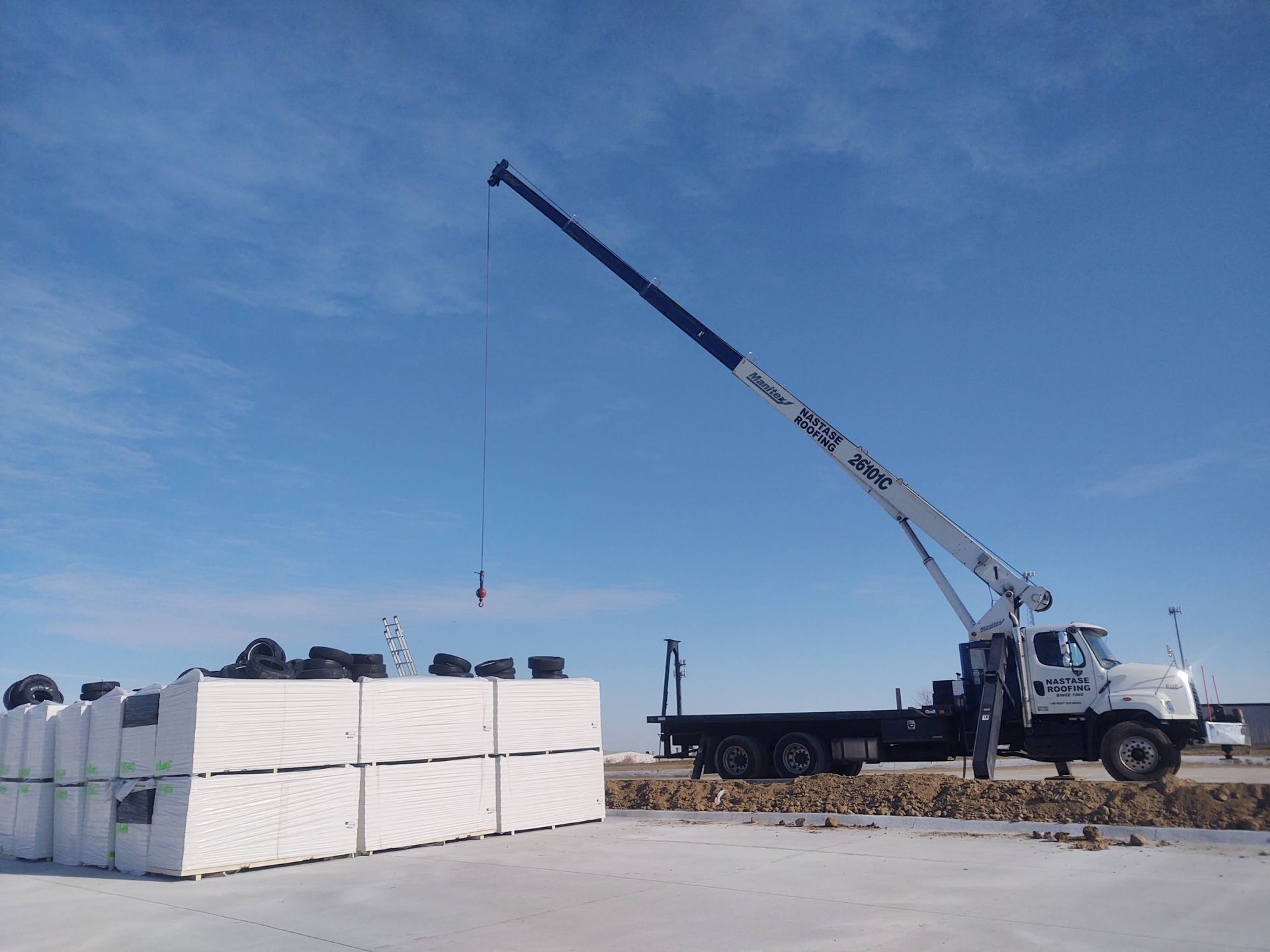 A white crane truck on a dirt lot beside a large stack of white rectangular building materials under a blue sky.